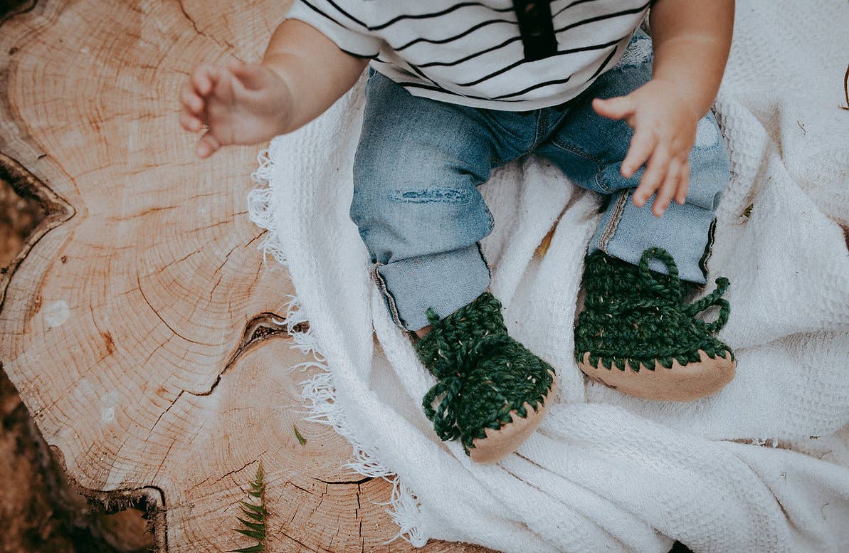 
                  
                    A baby's legs and hands are visible as they sit on a white fringed blanket on a rustic wood log. The baby is wearing a black and white striped shirt, blue jeans, and dark green knitted booties with tan soles.
                  
                