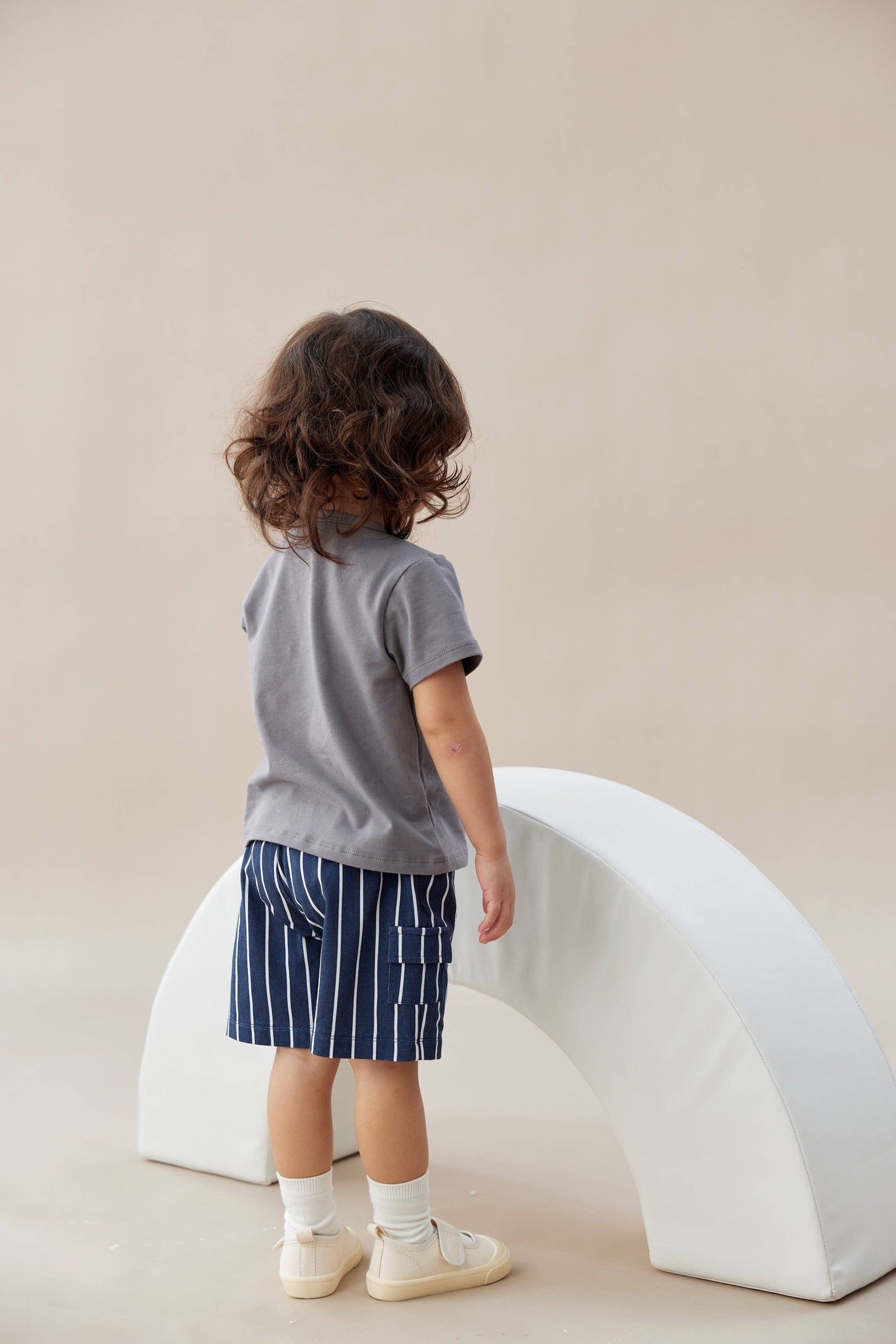 
                  
                    Child wearing a gray shirt and blue striped shorts standing in front of a white arch on a beige background.
                  
                