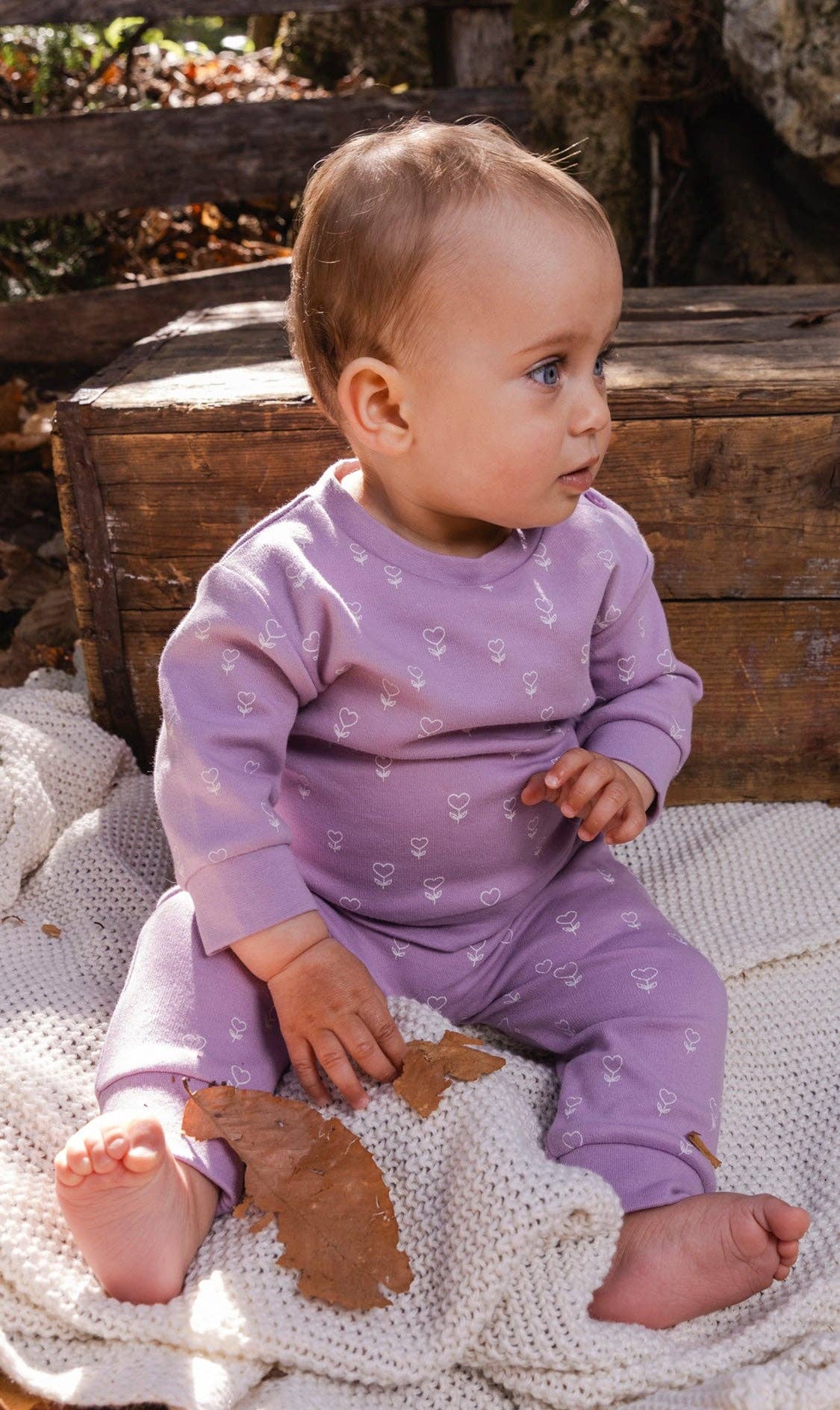 
                  
                    A baby sitting outdoors on a cream knit blanket, wearing a light purple long-sleeve top and pant set with a small white pattern..
                  
                