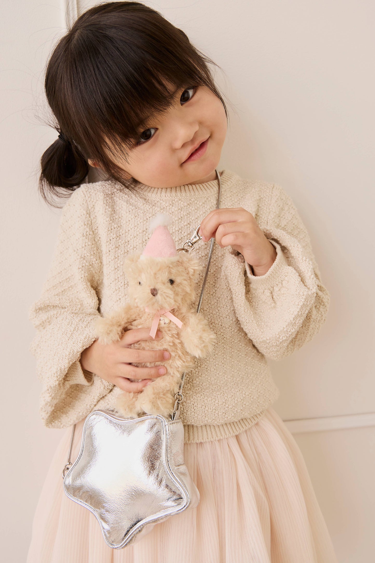 
                  
                    Young girl holding a teddy bear and a silver star-shaped bag against a white background.
                  
                