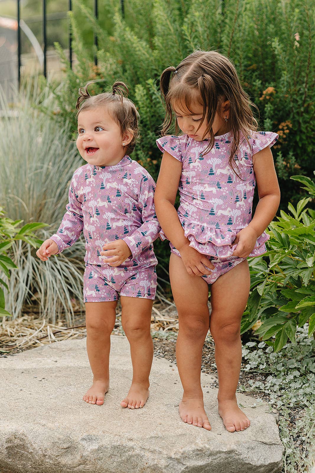 
                  
                    Two young girls in matching pink swim outfits standing on a rock with greenery around.
                  
                