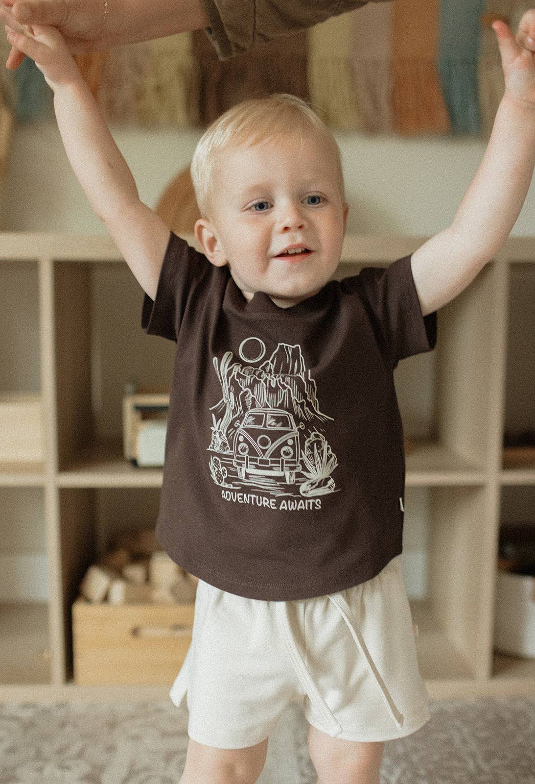 
                  
                    Child wearing a brown t-shirt with a graphic design, standing indoors with arms raised.
                  
                