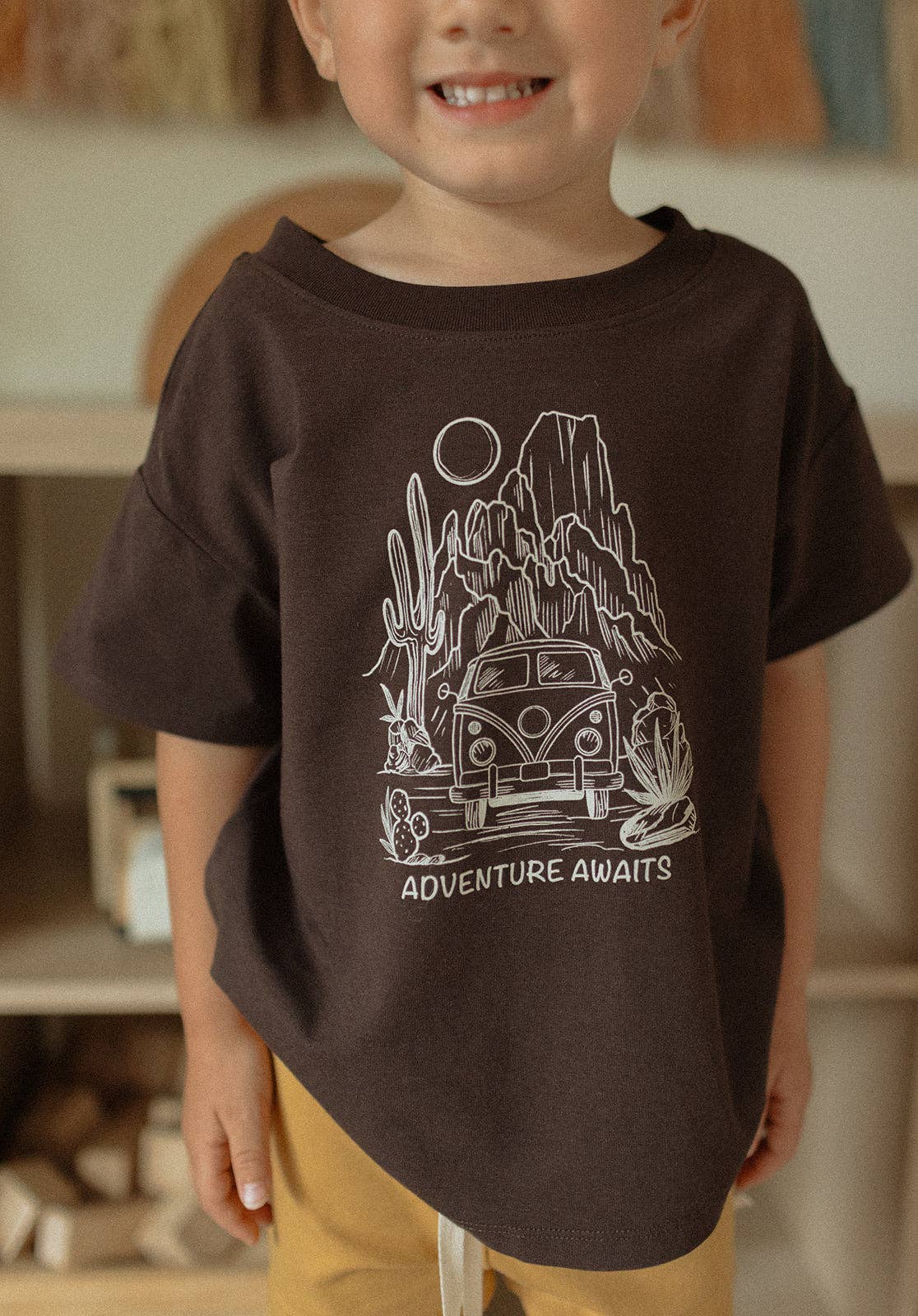 
                  
                    Child wearing a brown t-shirt with a graphic design of a van and desert landscape, text 'Adventure Awaits'.
                  
                