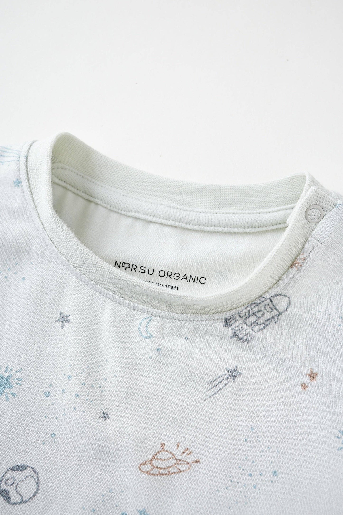 
                  
                    Close-up of a baby outfit with 'Norsu Organic' label and space-themed print on a white background.
                  
                