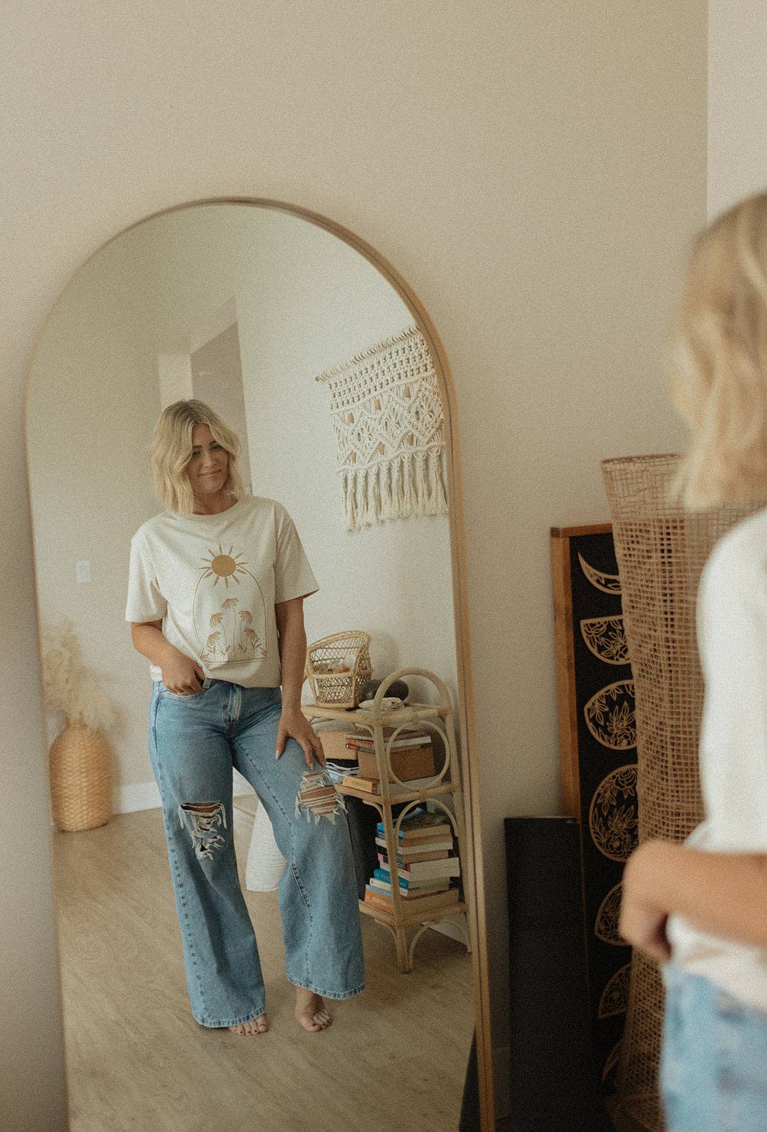 
                  
                    Woman standing in a room with a large mirror, wearing a white t-shirt and blue jeans.
                  
                