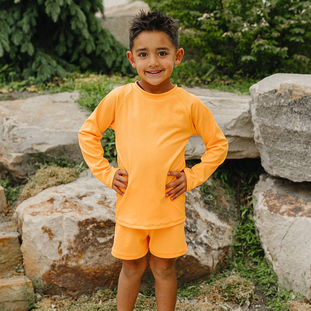 
                  
                    Child wearing an orange long-sleeve rash guard shirt and swim shorts standing outdoors with rocks and greenery in the background
                  
                