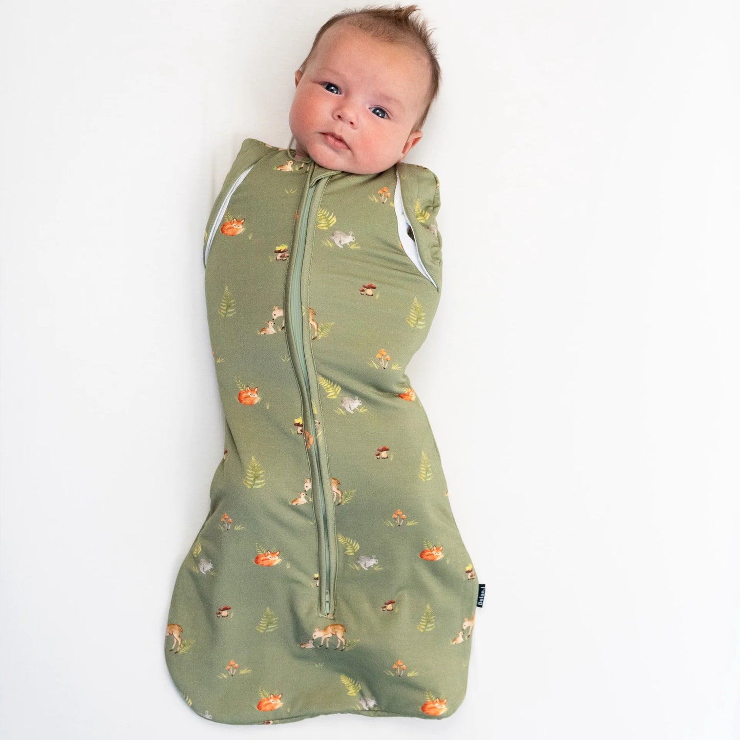 
                  
                    A baby wrapped in an olive green zippered swaddle blanket with a small woodland animal print, lying on a plain white background.
                  
                