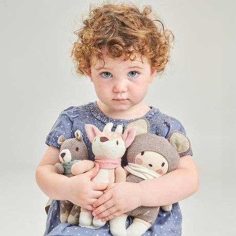 
                  
                    A toddler with curly light brown hair and blue eyes holds three soft plush toys against a plain light gray studio background. The child wears a dusty blue short-sleeve top, and the stuffed animals are in muted neutral tones with a pink scarf.
                  
                
