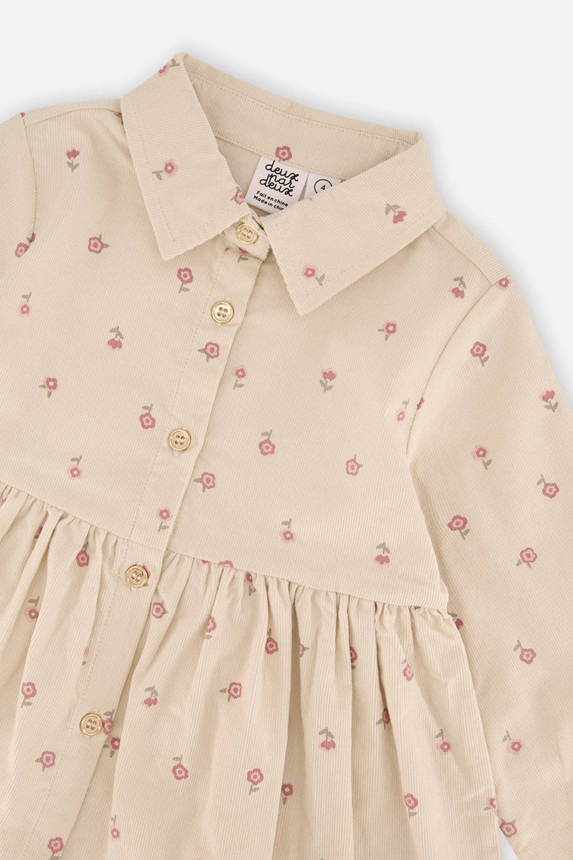 
                  
                    A close-up of a children's long-sleeve collared dress in a warm cream or light beige fabric, decorated with a small pink and green floral print. The dress features a buttoned front bodice with gold-tone buttons and a gathered waist seam.
                  
                