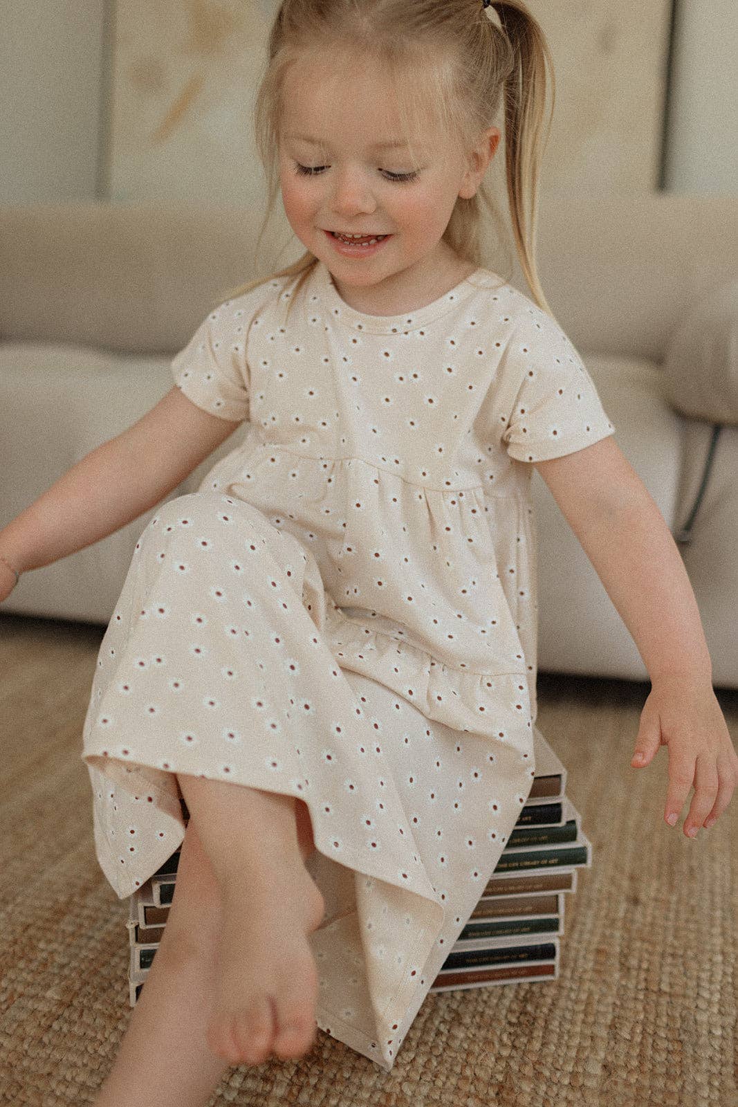 
                  
                    Young girl in a white dress with small patterns sitting on a stack of books in a living room.
                  
                