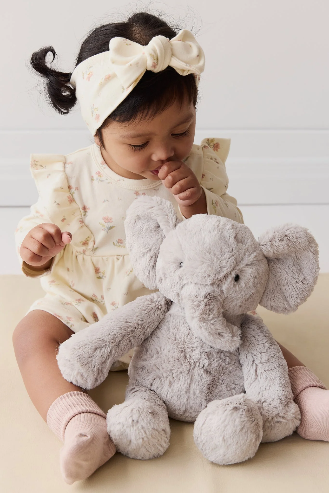 
                  
                    Baby sitting with a plush elephant toy wearing a white headband.
                  
                