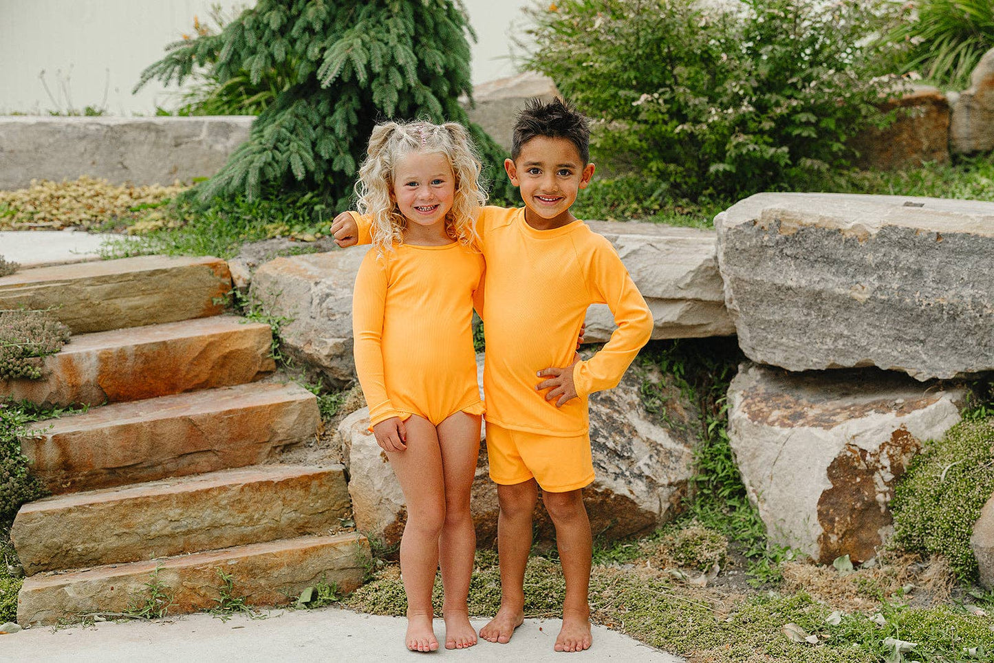 
                  
                    Two children in matching bright orange swim outfits standing on stone steps outdoors.
                  
                