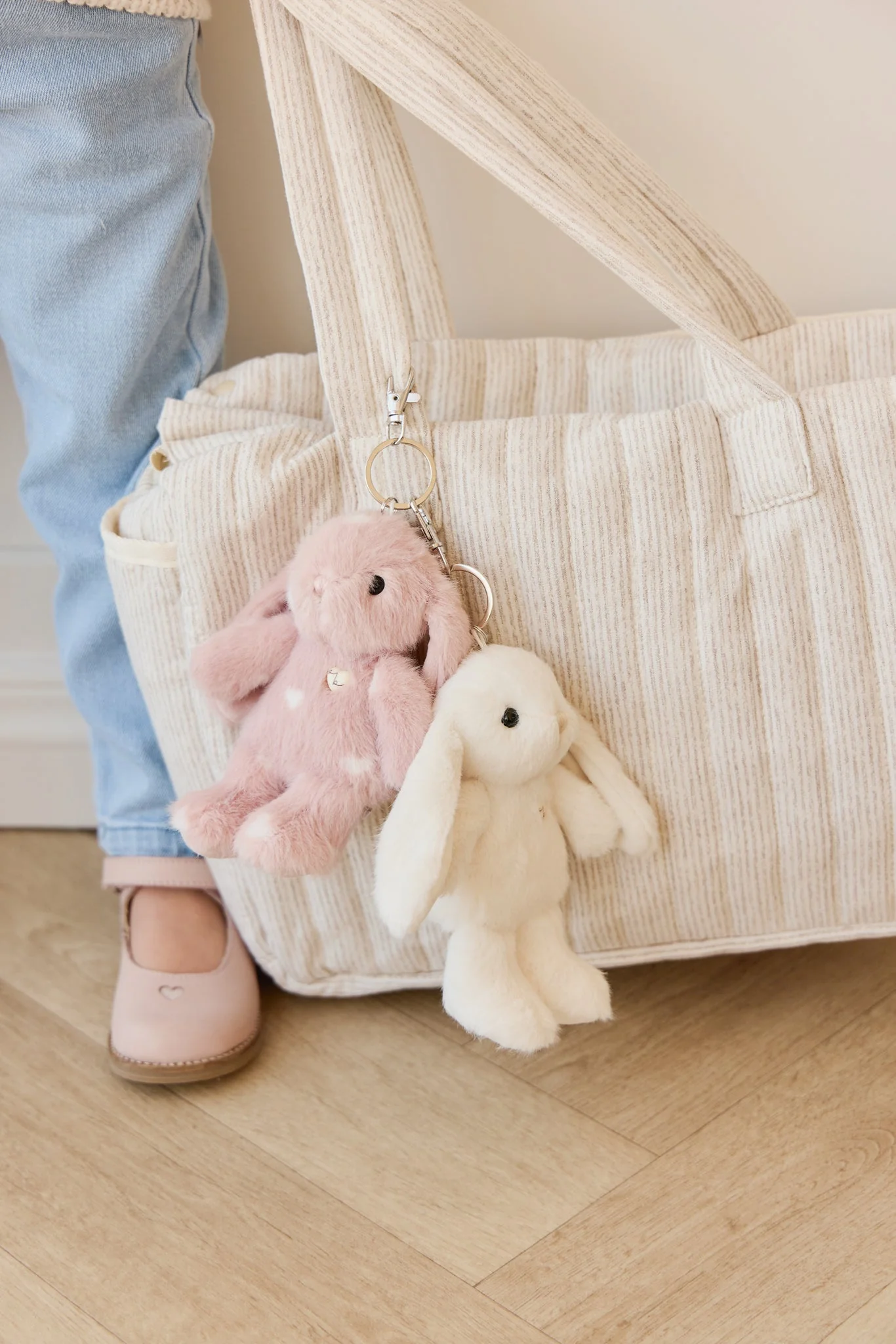 
                  
                    Close-up of a beige textured bag with plush toys attached, on a wooden floor.
                  
                