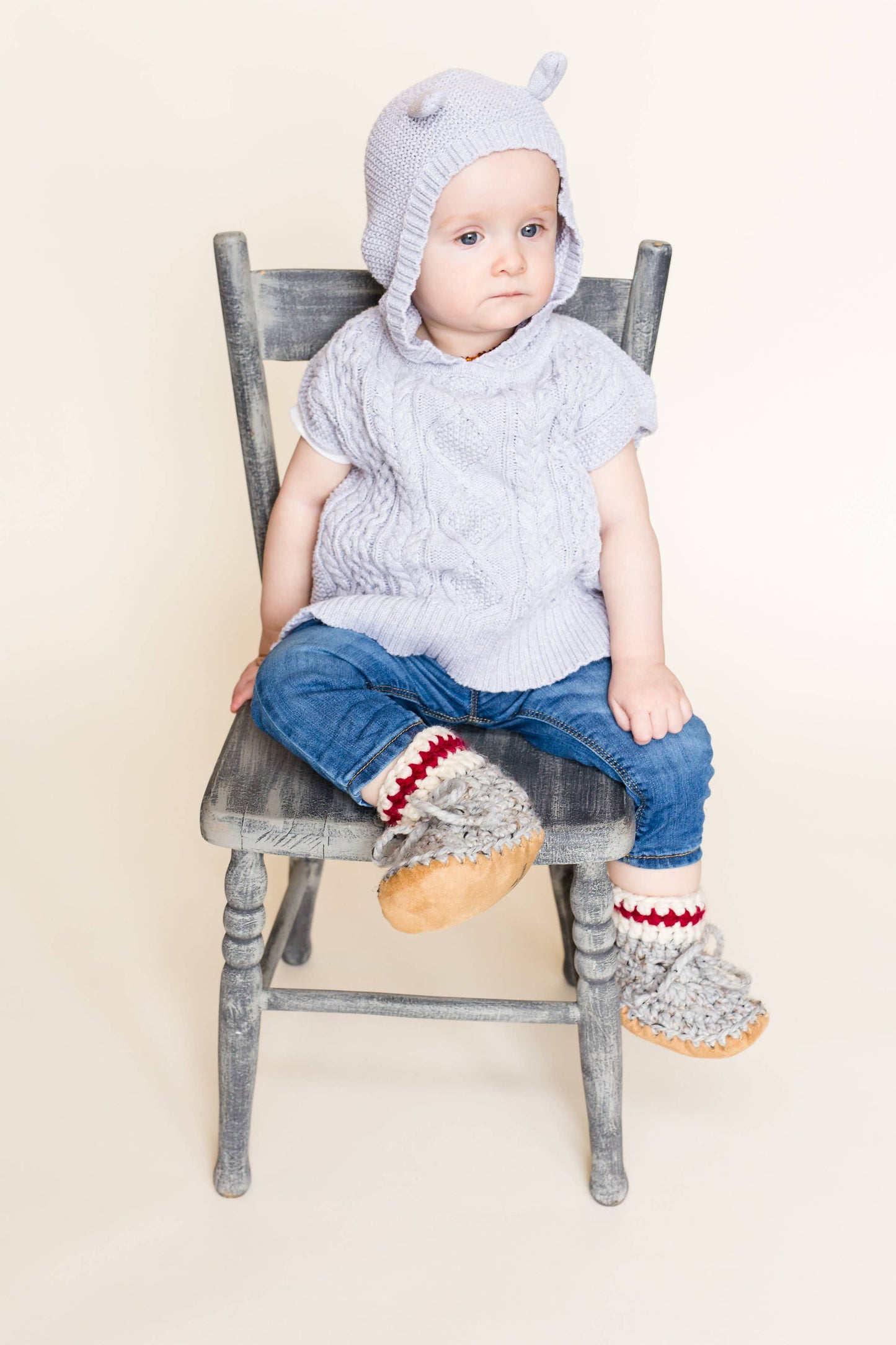
                  
                    Child sitting on a wooden chair wearing a knit sweater, blue jeans, and grey wool booties against a white background. 
                  
                