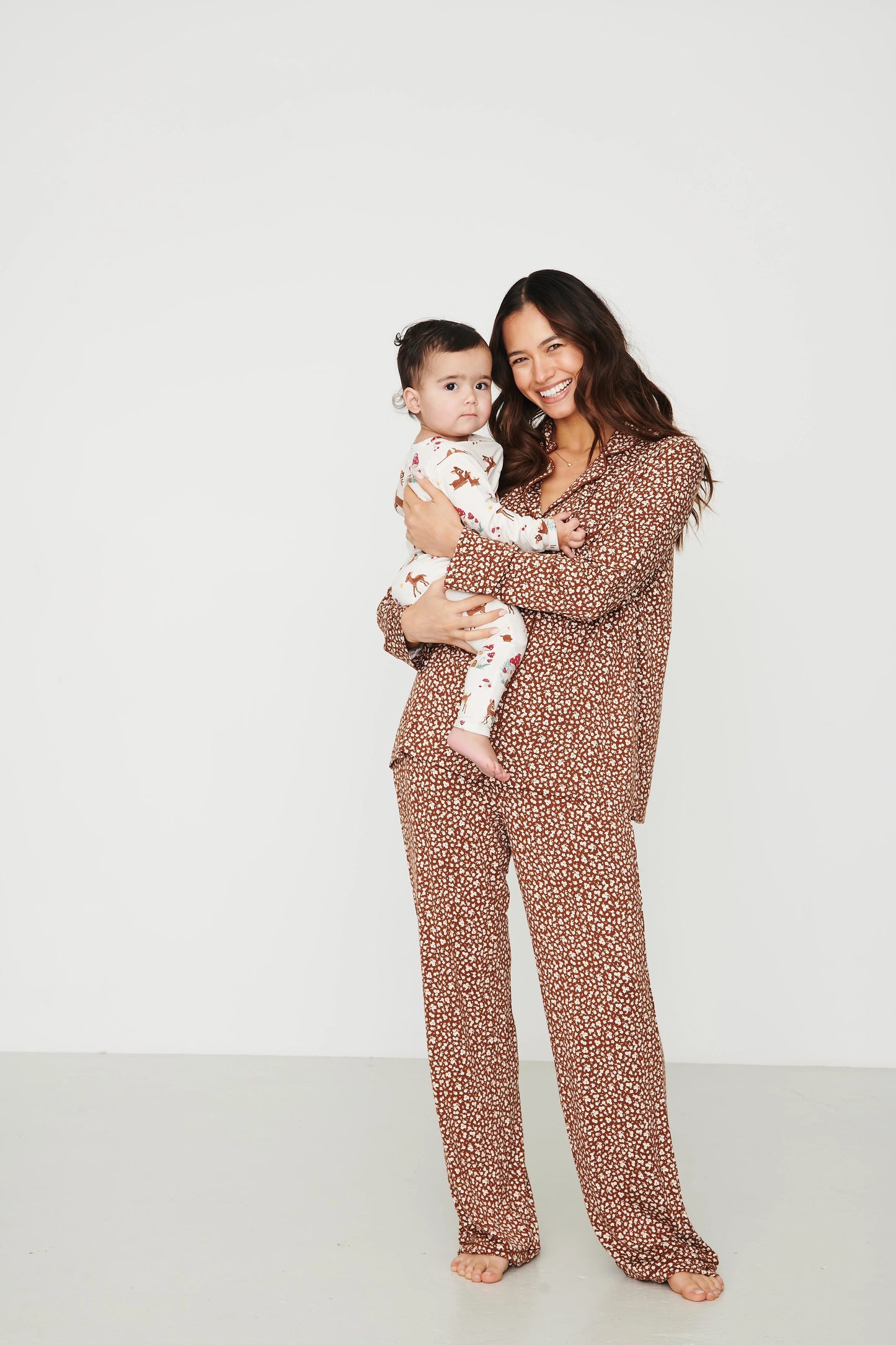 
                  
                    A woman holding a baby wearing matching two-piece pajamas with a brown and beige animal print pattern, set against a plain light gray studio background.
                  
                