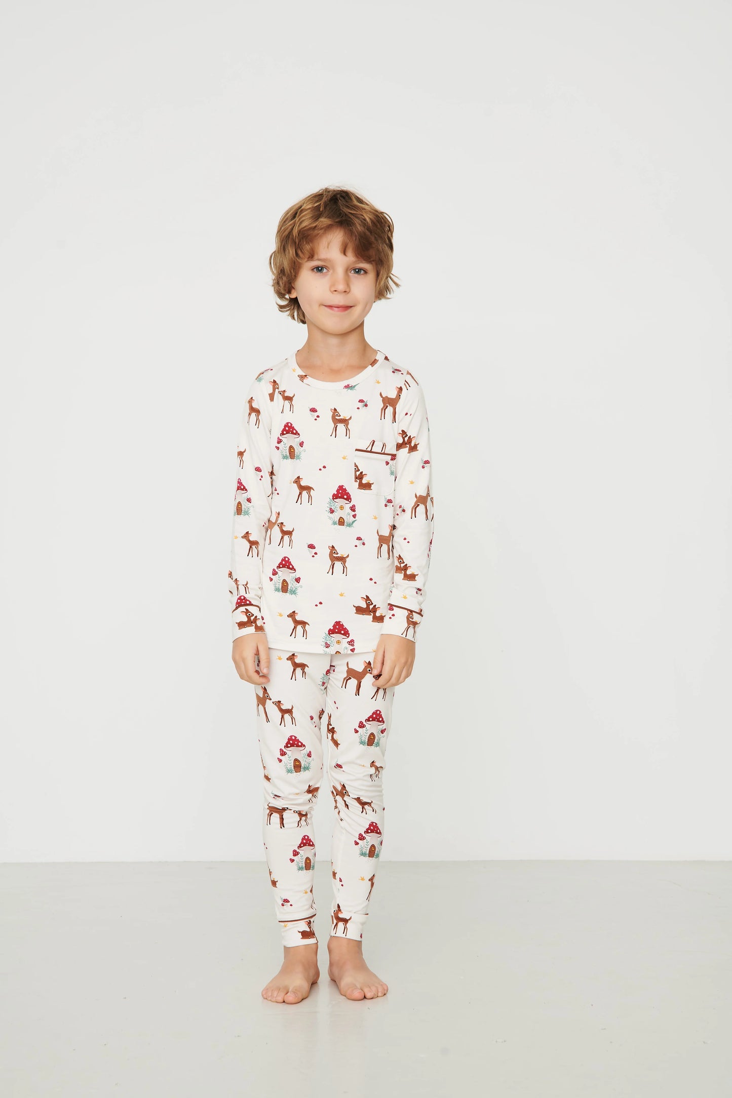 
                  
                    A child stands barefoot against a plain white studio background, wearing a long-sleeve pajama top and matching pants with an all-over deer print.
                  
                