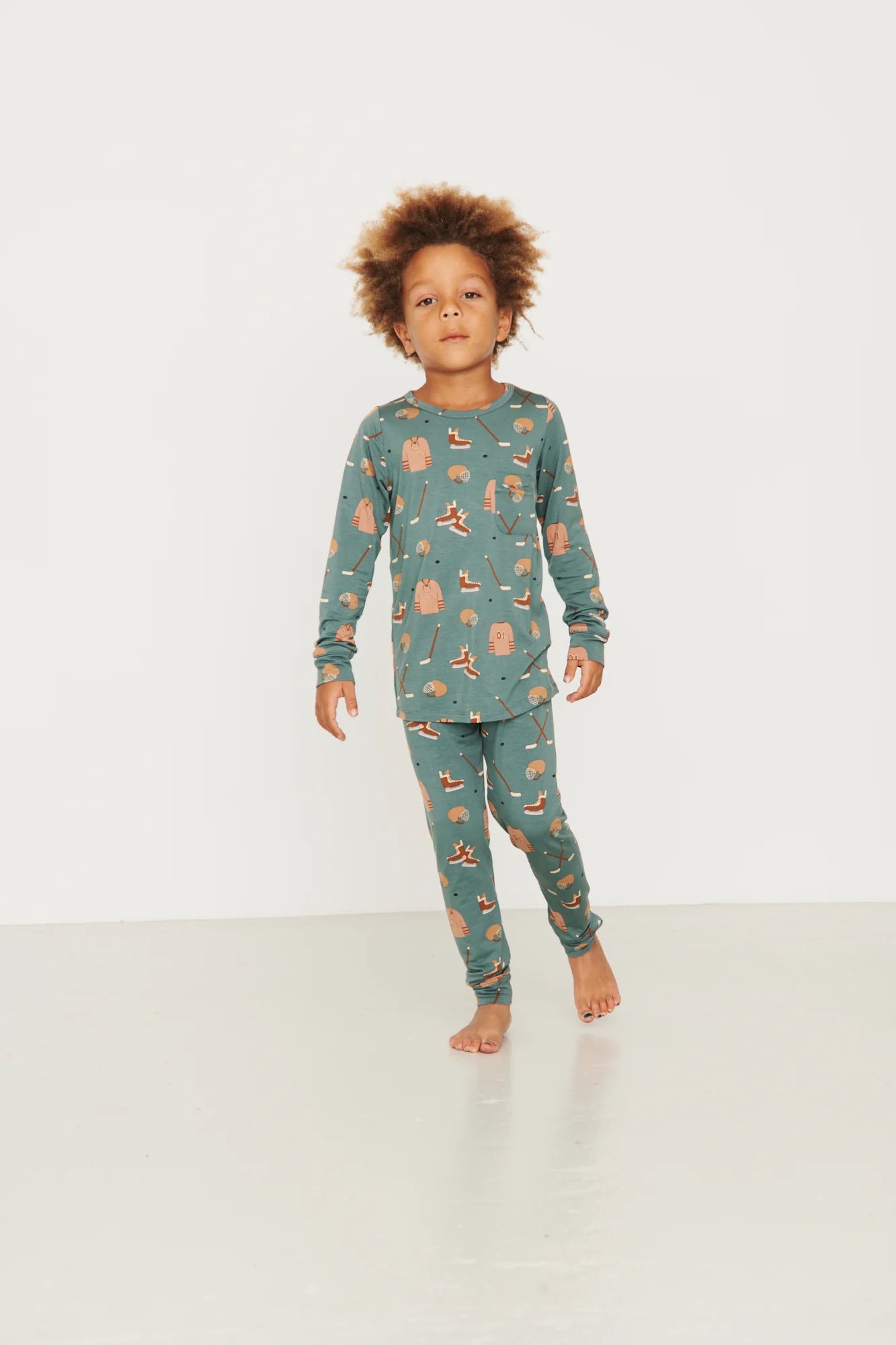
                  
                    A young child with curly brown hair stands barefoot in a studio wearing a long-sleeve pajama set in a muted teal-green color with a small orange and beige hockey print.
                  
                