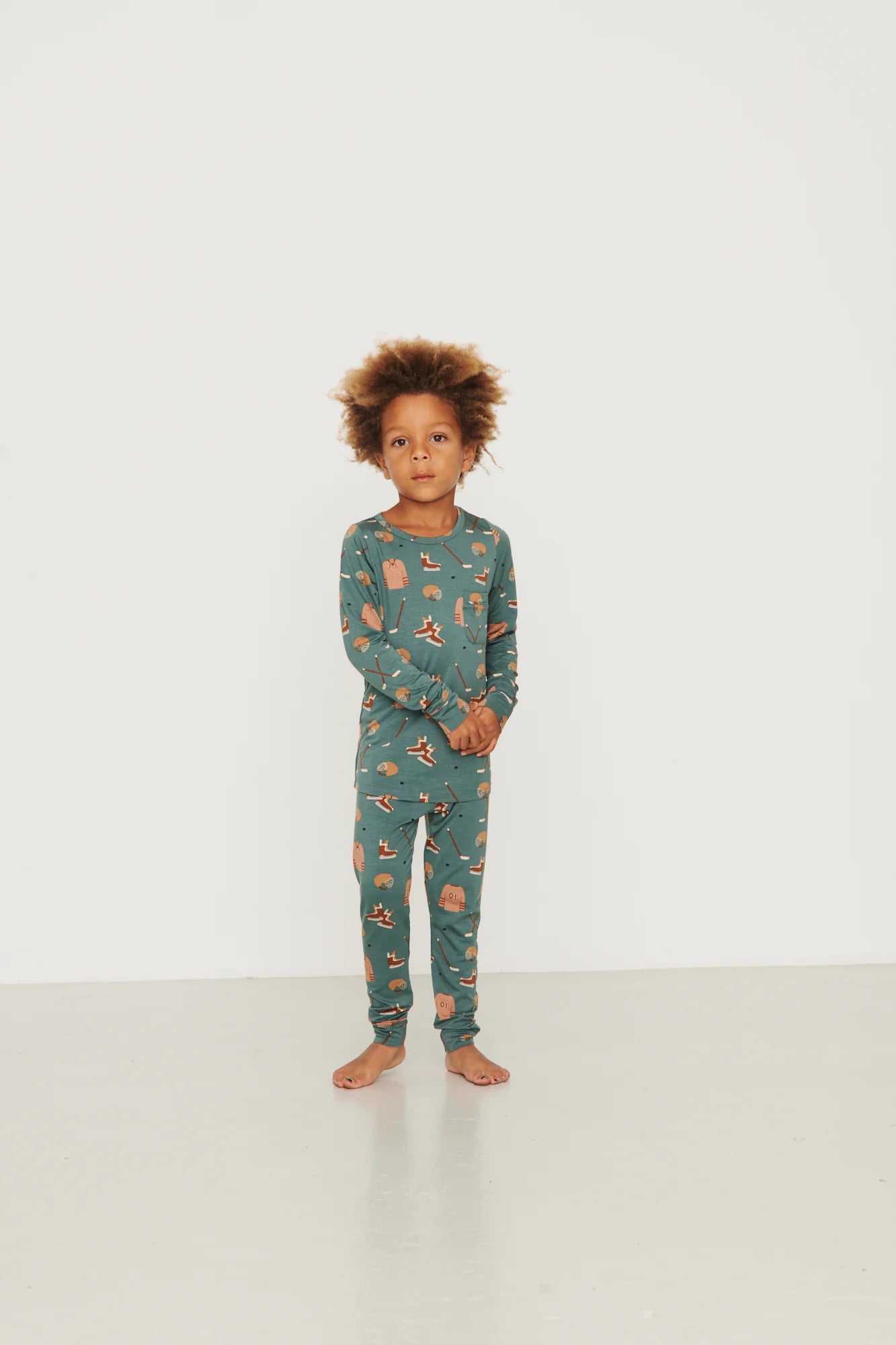 
                  
                    A young child with curly brown hair stands barefoot in a studio setting, wearing a long-sleeve pajama set in a muted teal-green color with a small all-over pattern of an orange and beige hockey print.
                  
                