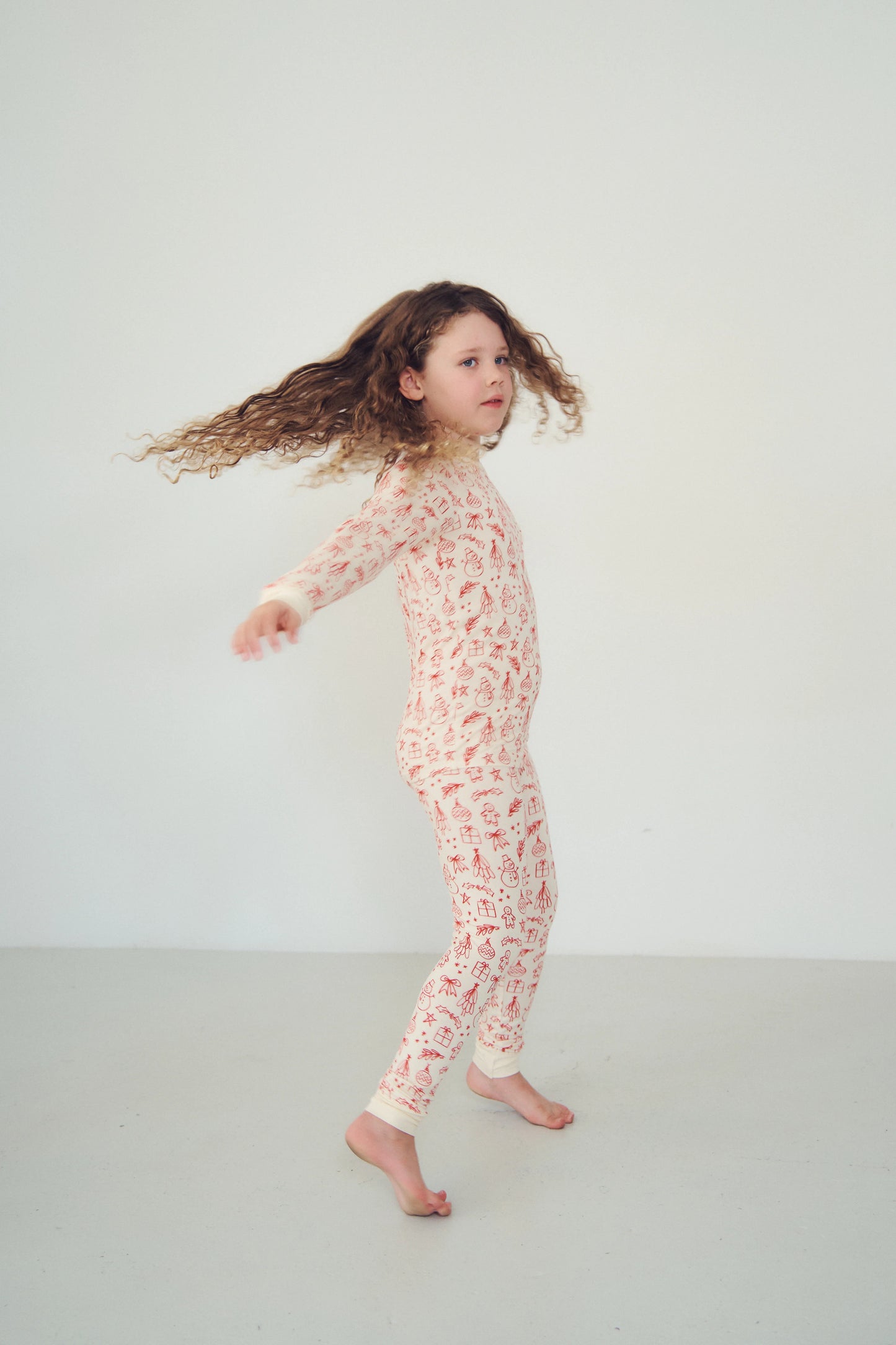 
                  
                    A young girl with long, curly brown hair is captured mid-motion while wearing a cream-colored two-piece pajama set with a red Christmas pattern, set against a plain light gray studio background.
                  
                