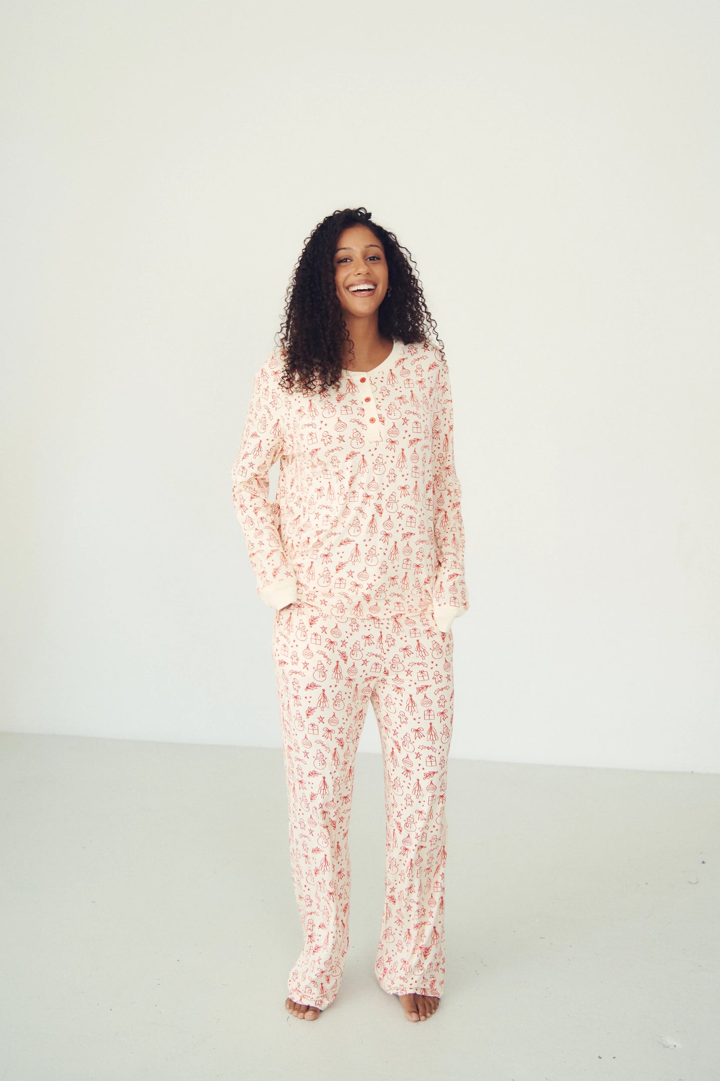 
                  
                    A woman stands barefoot in a studio wearing a matching long-sleeve pajama set with a Christmas print. The pajamas are a light peach-pink base with a dense red and white pattern.
                  
                