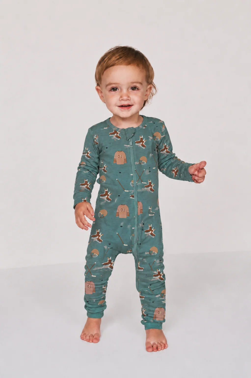 A toddler standing and smiling while wearing a long-sleeve footless pajama one-piece in a muted teal-green color with a repeating hockey print in brown and tan, photographed against a plain light gray studio background.