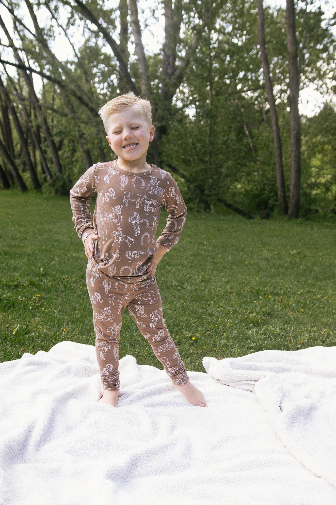 
                  
                    Child wearing a brown pajama set with a rodeo print standing on a blanket in a grassy area with trees.
                  
                