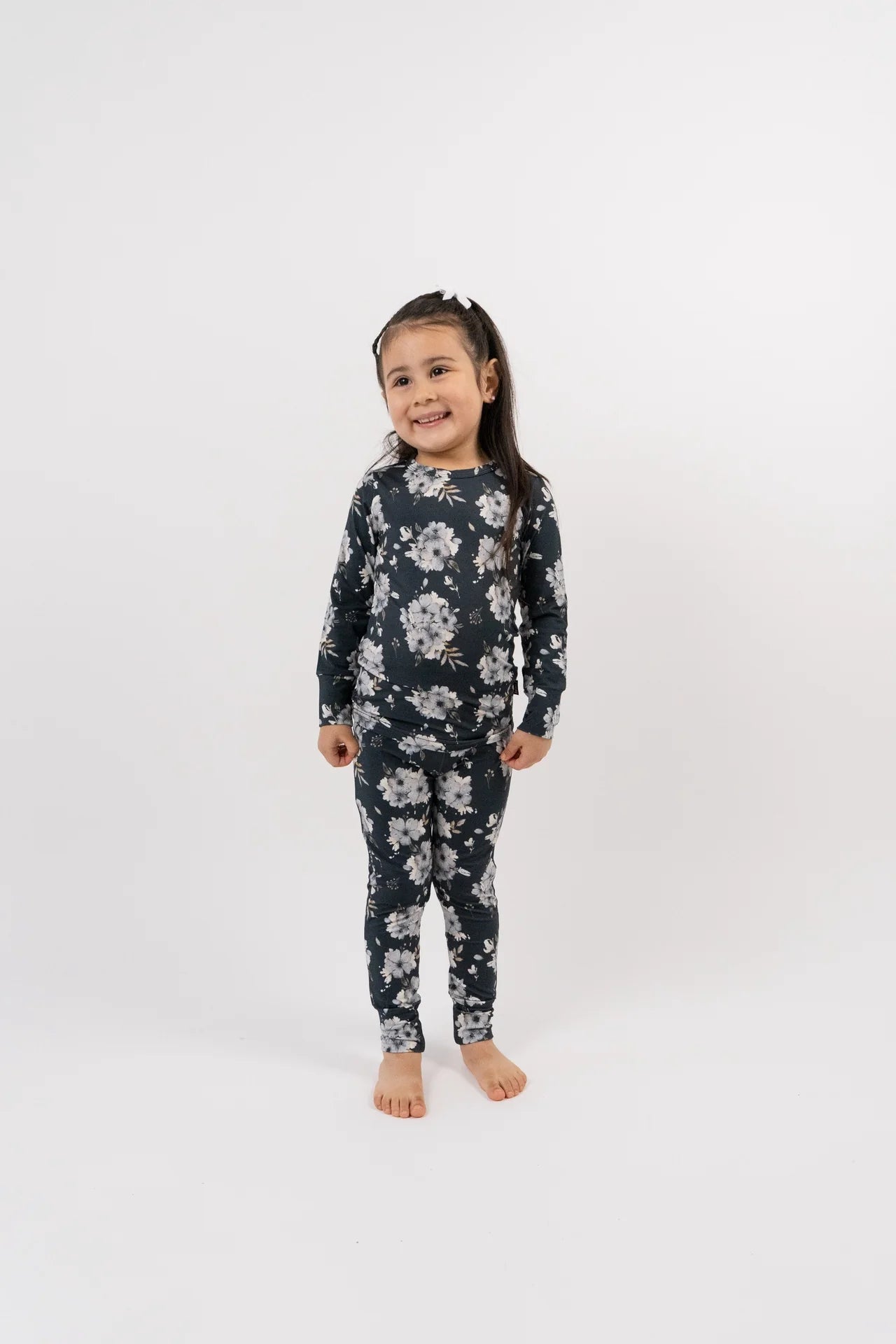 
                  
                    A young girl stands barefoot against a plain white studio background, wearing a long-sleeve one-piece pajama set with a dark navy or black base and a light gray floral pattern.
                  
                