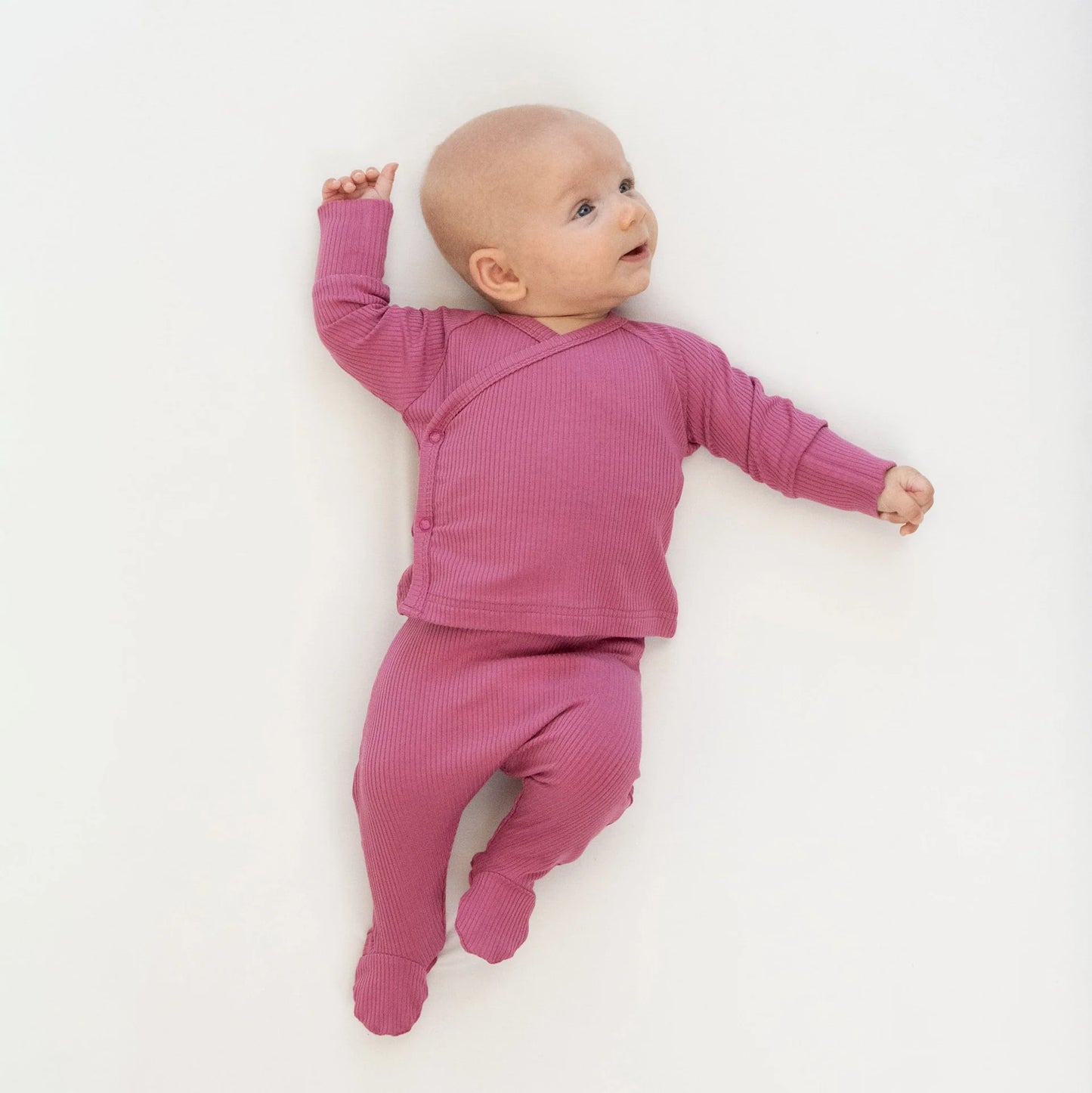 
                  
                    A baby lying on their back wearing a dusty pink ribbed knit footed sleeper with a snap-button closure, photographed against a clean white background.
                  
                