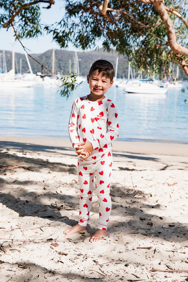 Child in red heart-patterned pajamas standing on a sandy beach with boats and trees in the background.
