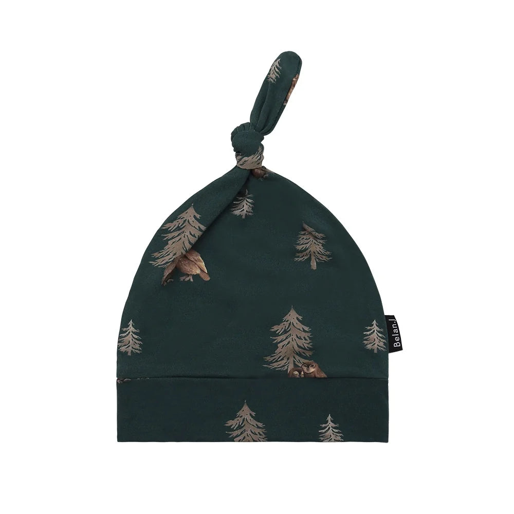 
                  
                    A dark forest green baby beanie hat featuring a repeating pattern of pine trees and owls, with a matching knotted strap at the top and a small black brand tag on the side.
                  
                