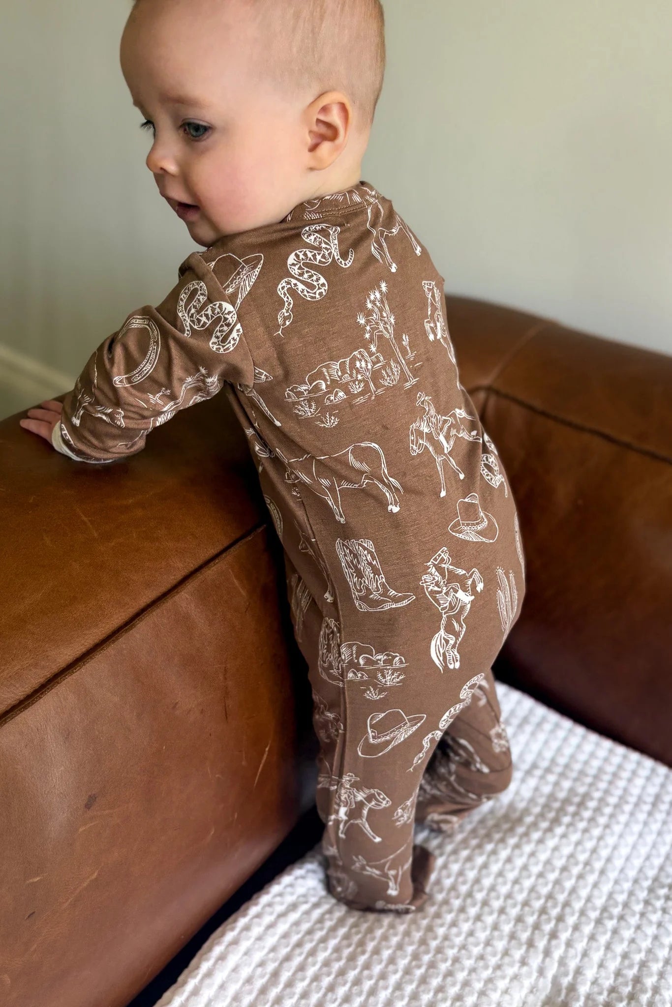 
                  
                    Baby in a brown rodeo patterned onesie standing on a couch.
                  
                