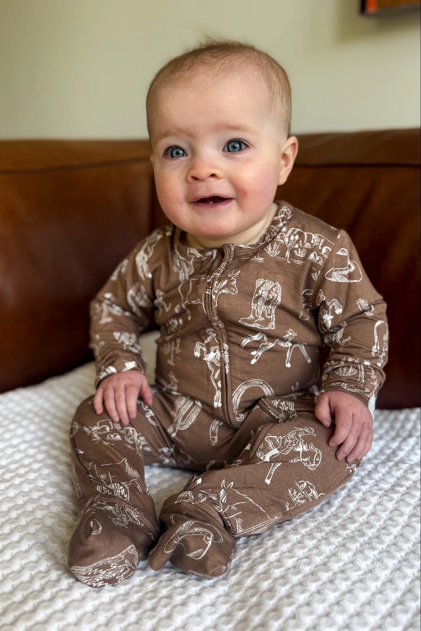 
                  
                    Baby wearing a brown rodeo patterned onesie sitting on a couch.
                  
                