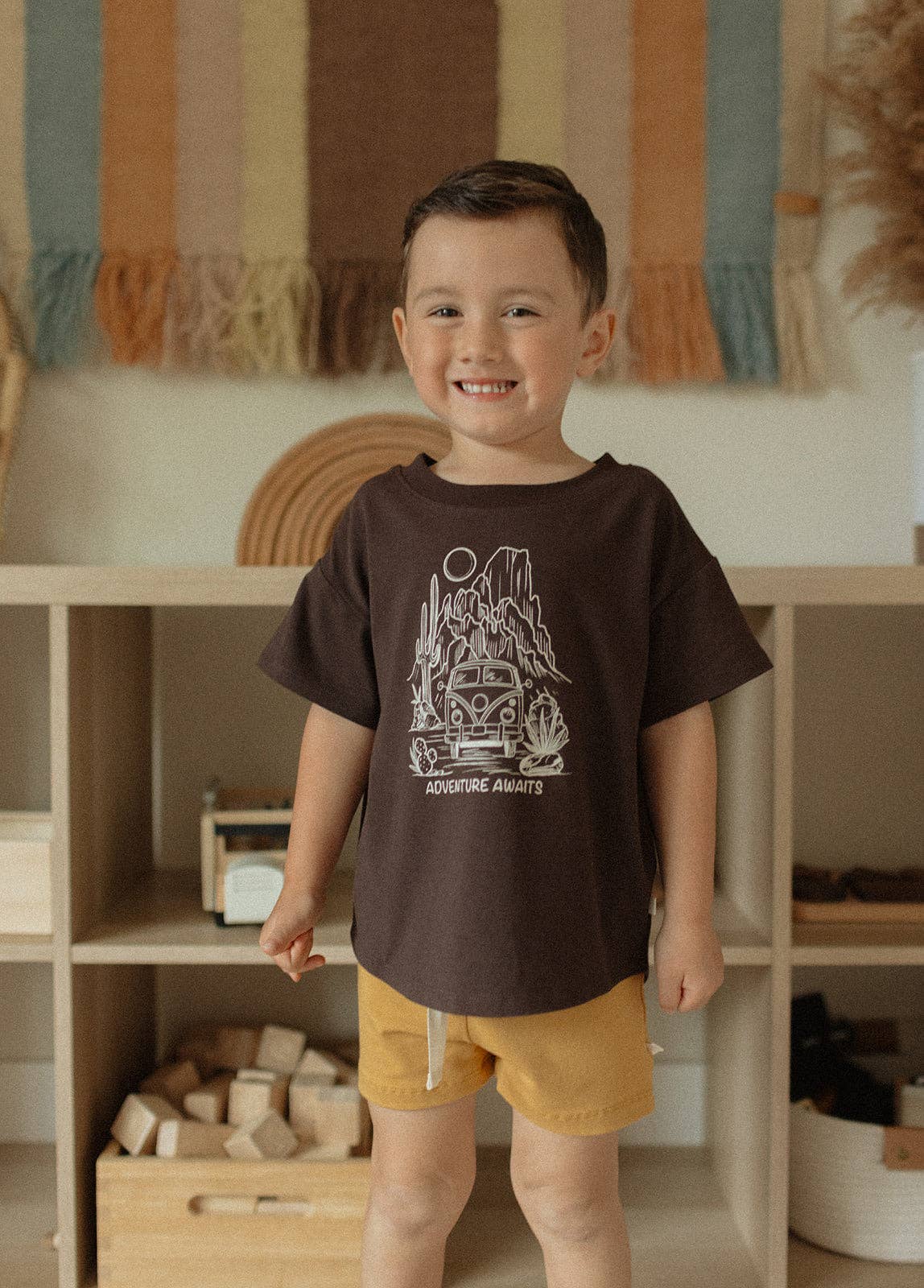 
                  
                    Child wearing a brown t-shirt with a graphic design, standing in a room with shelves and toys.
                  
                