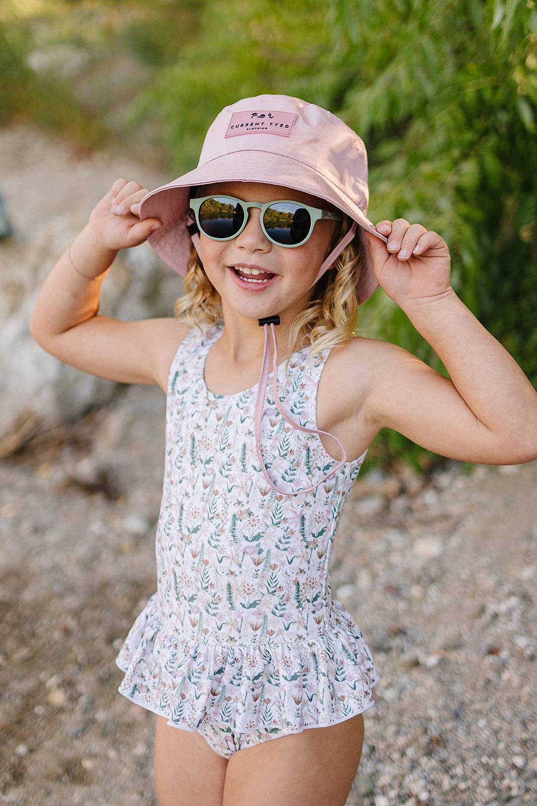 
                  
                    Young girl in a floral swimsuit and pink hat with sunglasses on a natural background.
                  
                