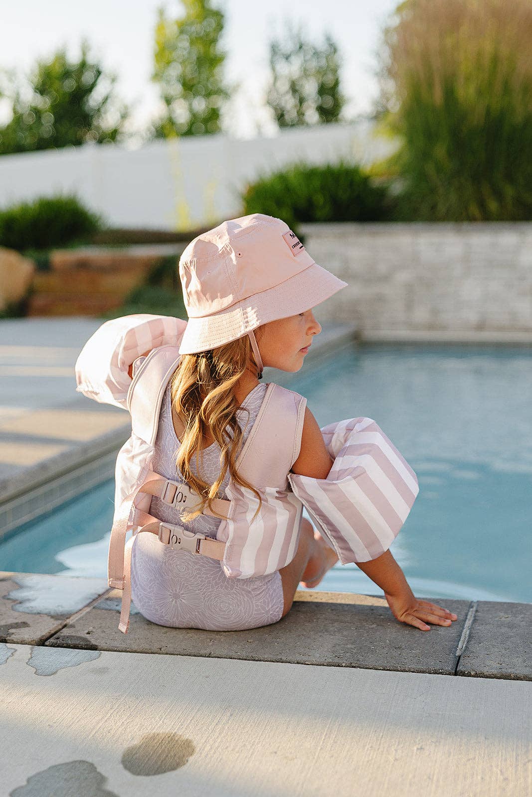 
                  
                    Child in a pink hat and outfit sitting by a pool with a blurred garden background.
                  
                