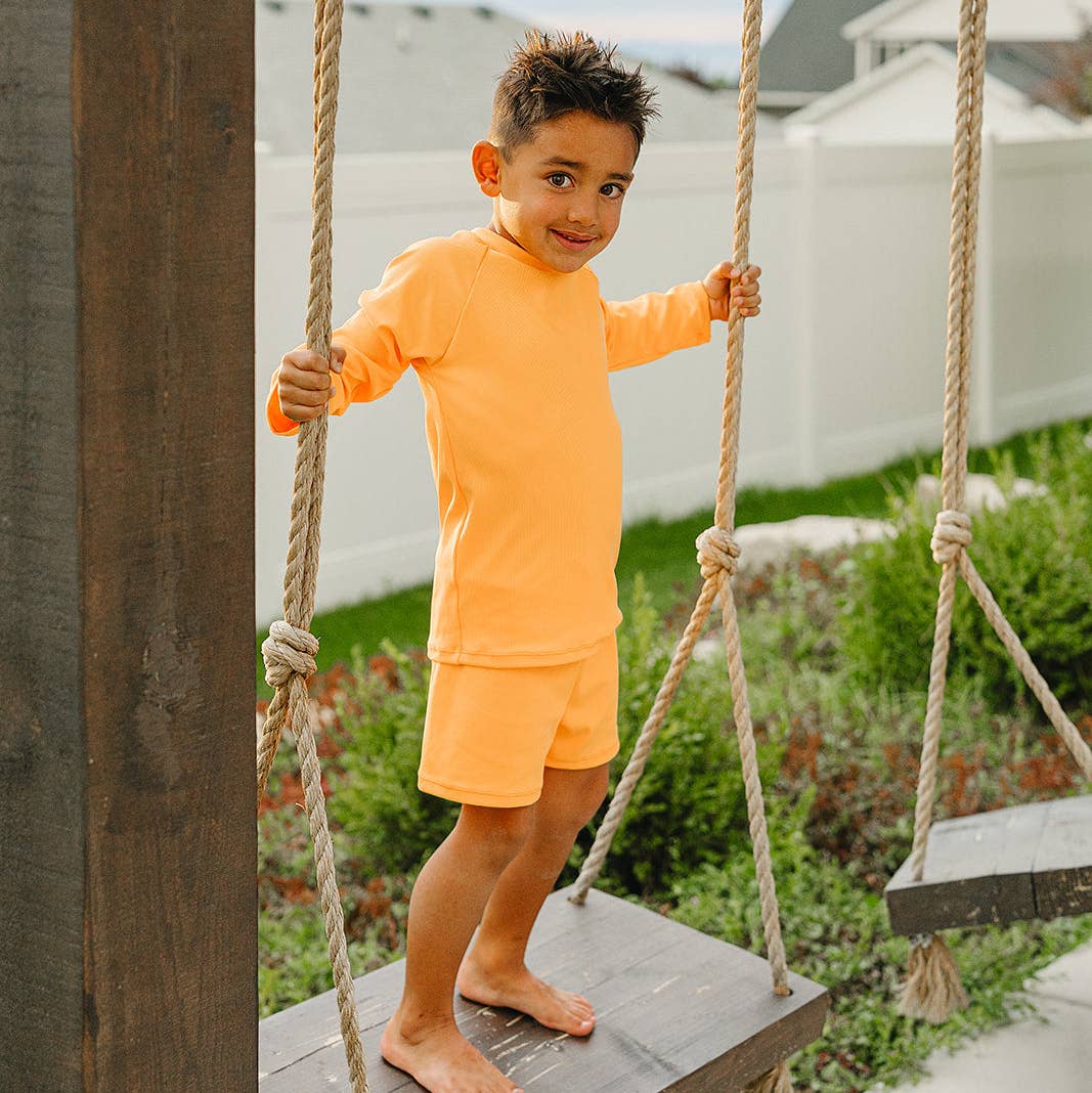 
                  
                    Child in orange swim outfit standing on a wooden swing set outdoors
                  
                