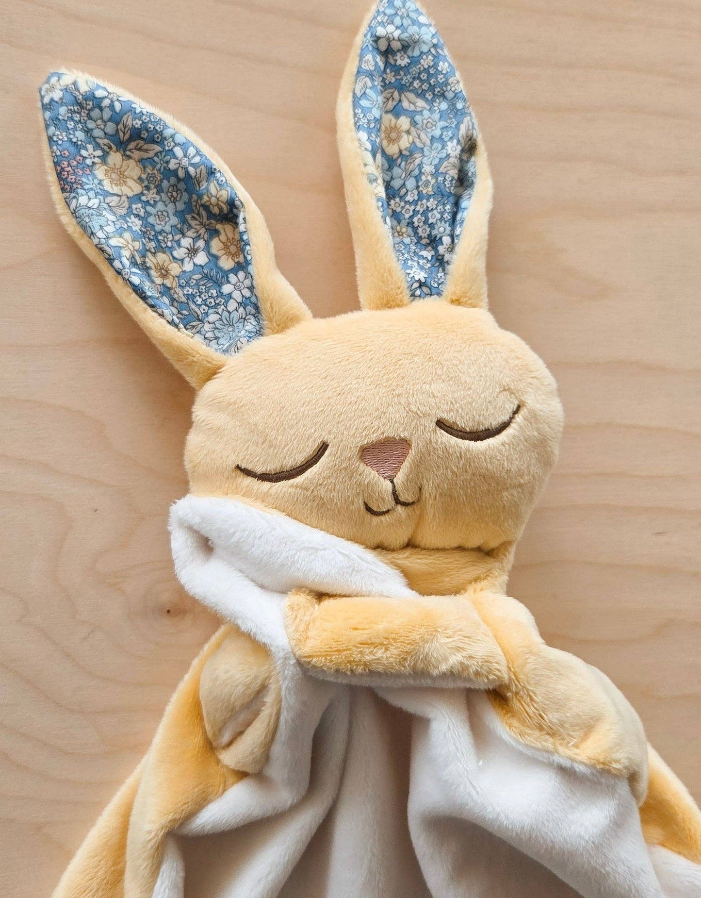 
                  
                    Plush rabbit toy with floral ears and a white blanket on a wooden background.
                  
                
