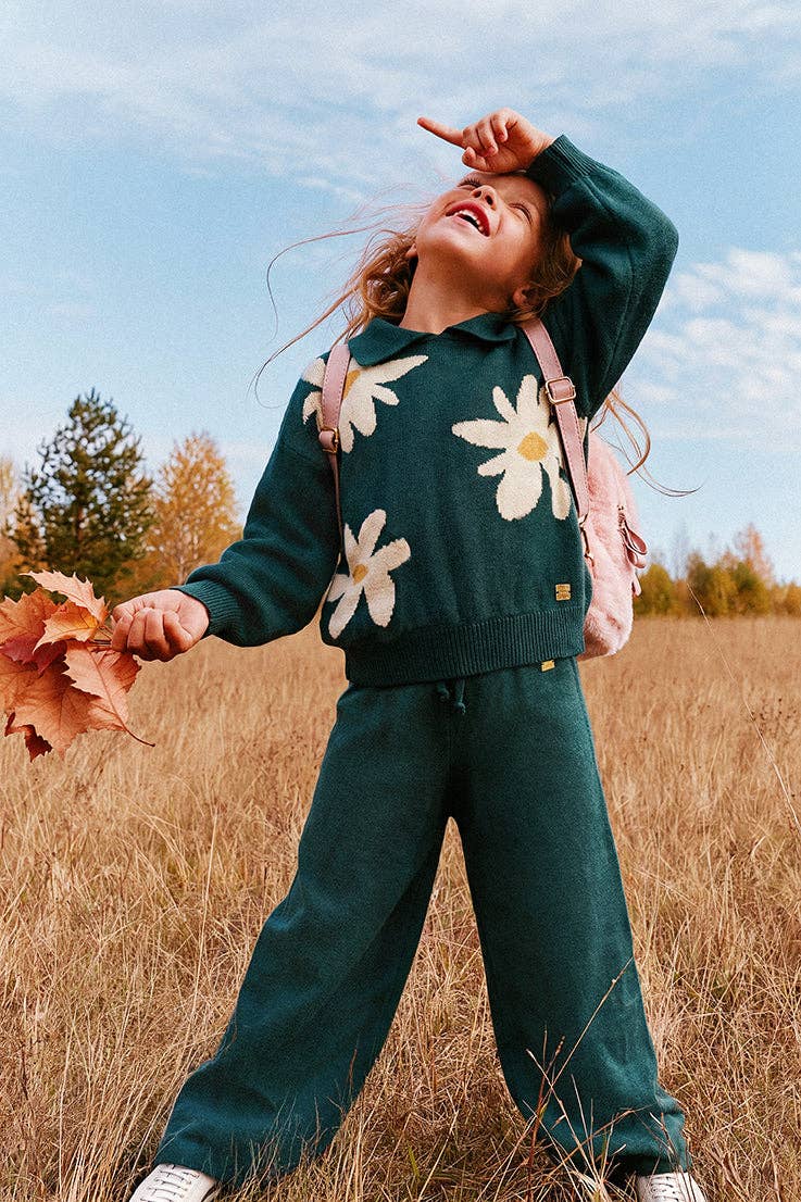 
                  
                    A young girl wearing a dark green knit matching set with large white daisy patterns on the sweater and pants, standing in a dry grass field under a blue sky.
                  
                