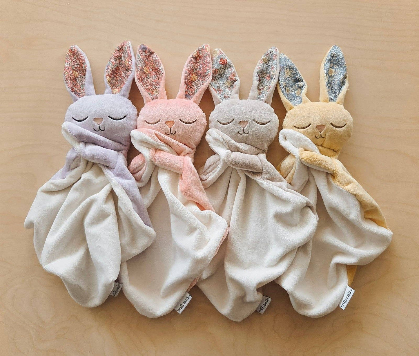 
                  
                    Set of four soft toy rabbit blankets in different colors on a wooden surface.
                  
                
