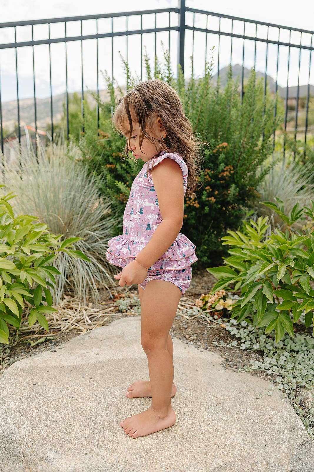 
                  
                    Young girl in a floral swim suit standing on a stone path with greenery around.
                  
                