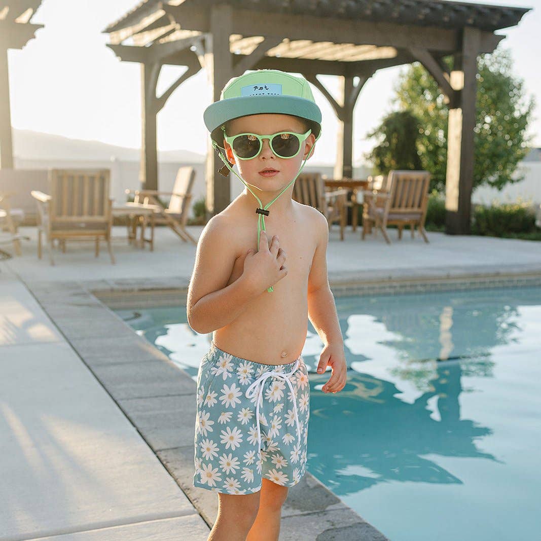 Child wearing a cap, sunglasses, and floral swim shorts by a poolside.