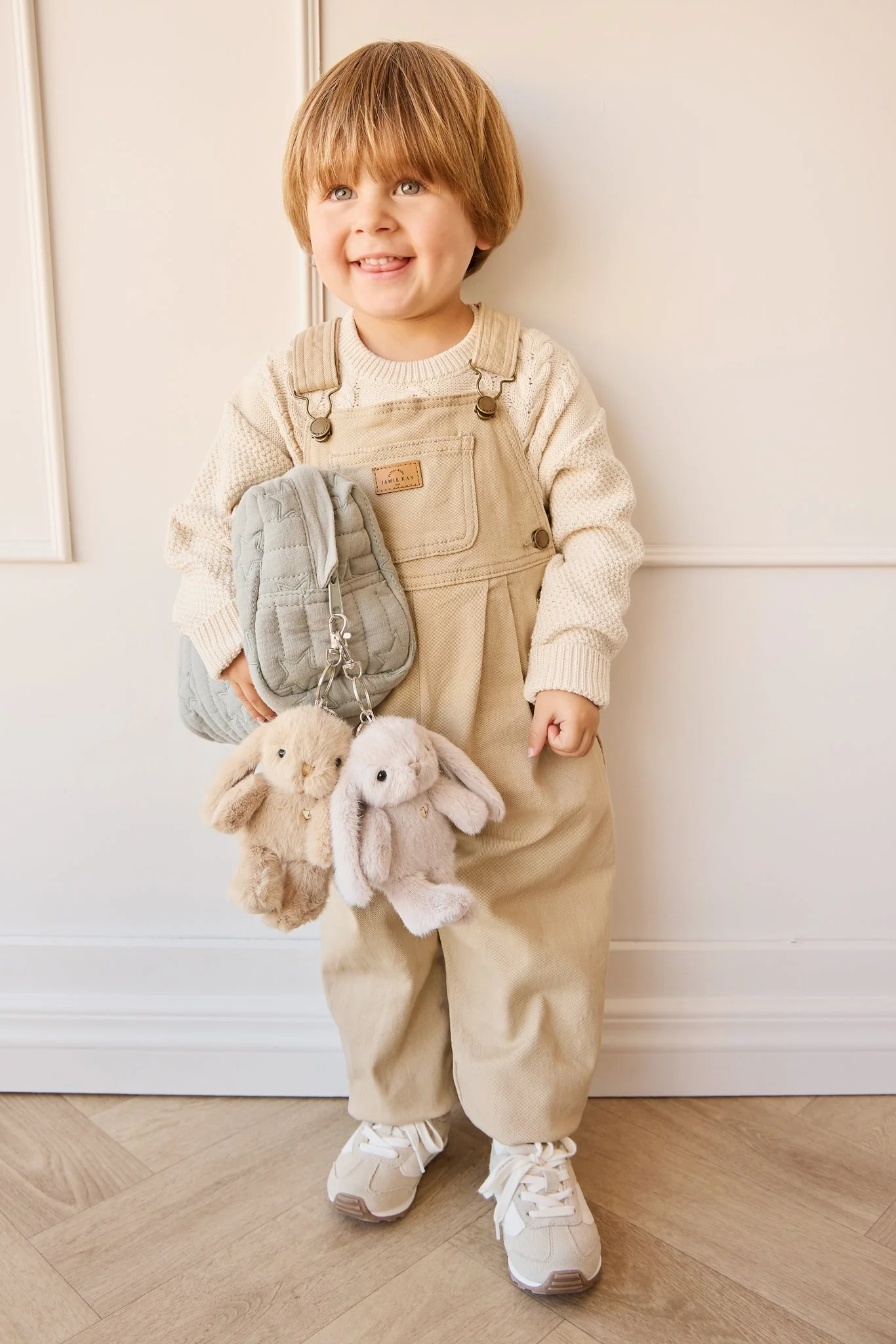 
                  
                    Child wearing beige overalls holding plush bunny toys against a neutral background.
                  
                