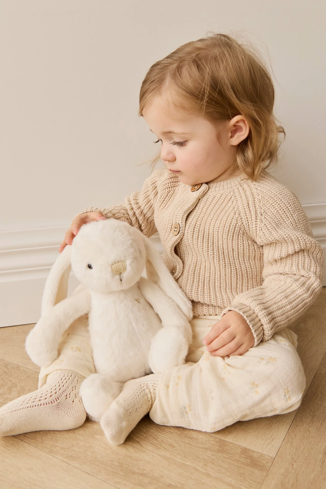 
                  
                    Child sitting on a wooden floor holding a plush toy, wearing a beige knit cardigan and white pants.
                  
                