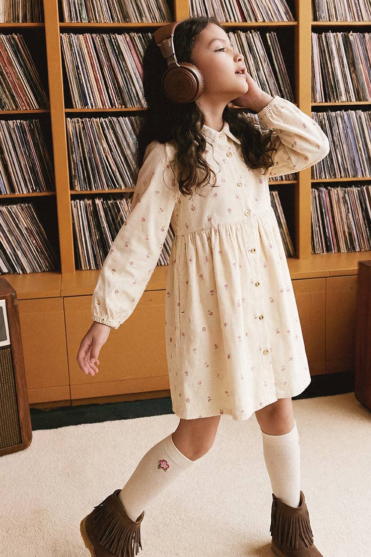 
                  
                    A young girl wearing a cream-colored long-sleeve dress with a tiny floral pattern, white knee-high socks, and brown fringe boots, standing in a room with wooden shelves filled with vinyl records.
                  
                
