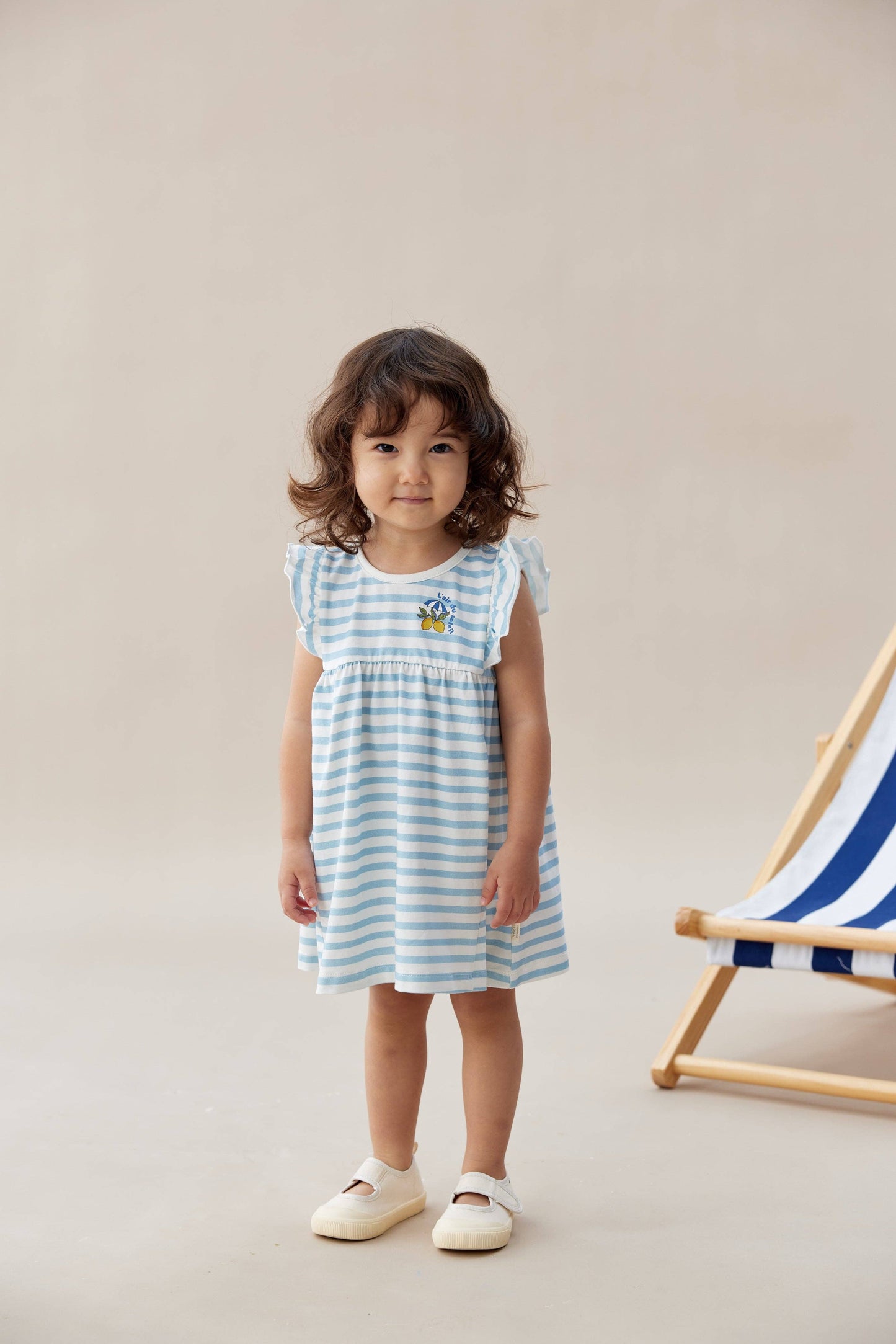Young girl wearing a blue and white striped dress standing next to a deck chair on a beige background.