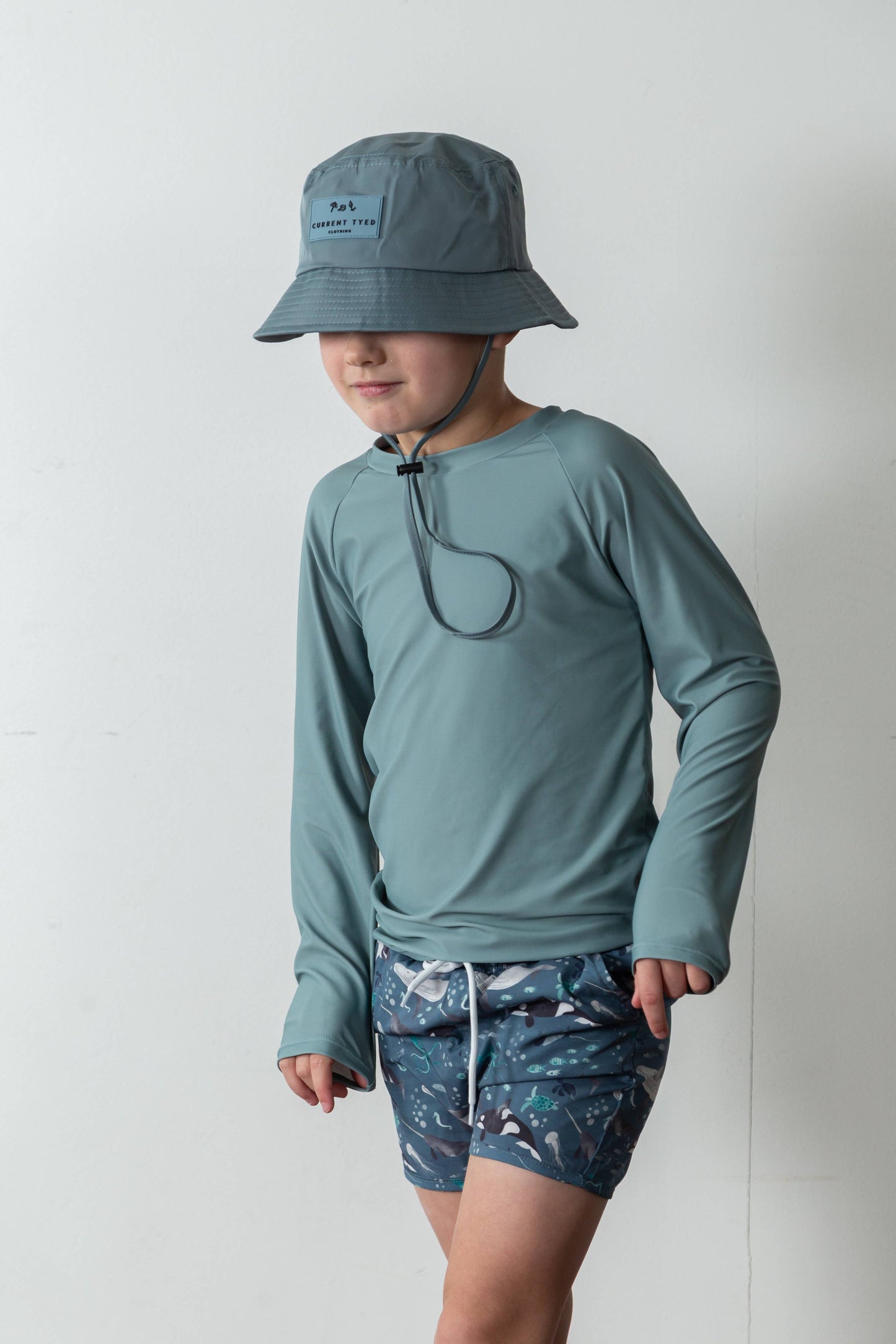 
                  
                    Child wearing a light blue long-sleeve shirt, bucket hat, and patterned shorts on a plain background.
                  
                