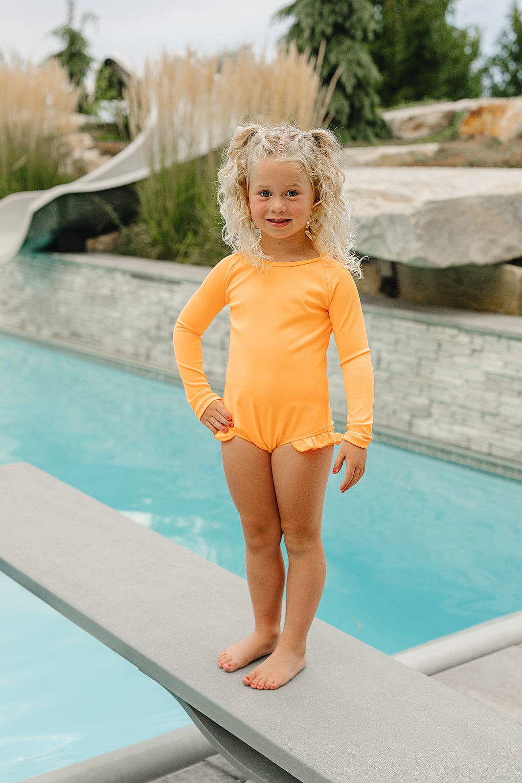 Child in an orange swimsuit standing by a pool with a scenic background