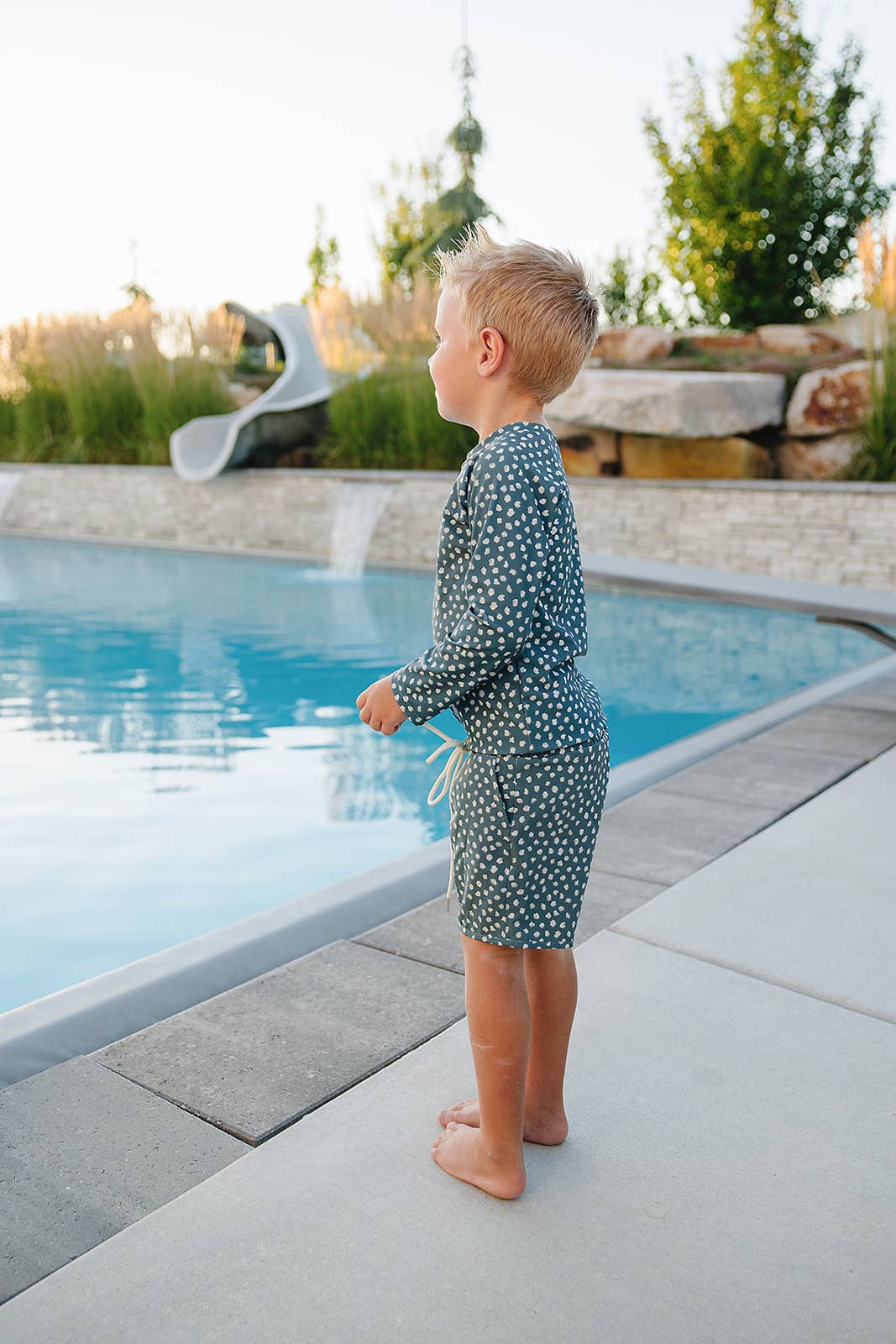 
                  
                    Child in a polka dot swimsuit standing by a pool with a scenic background
                  
                