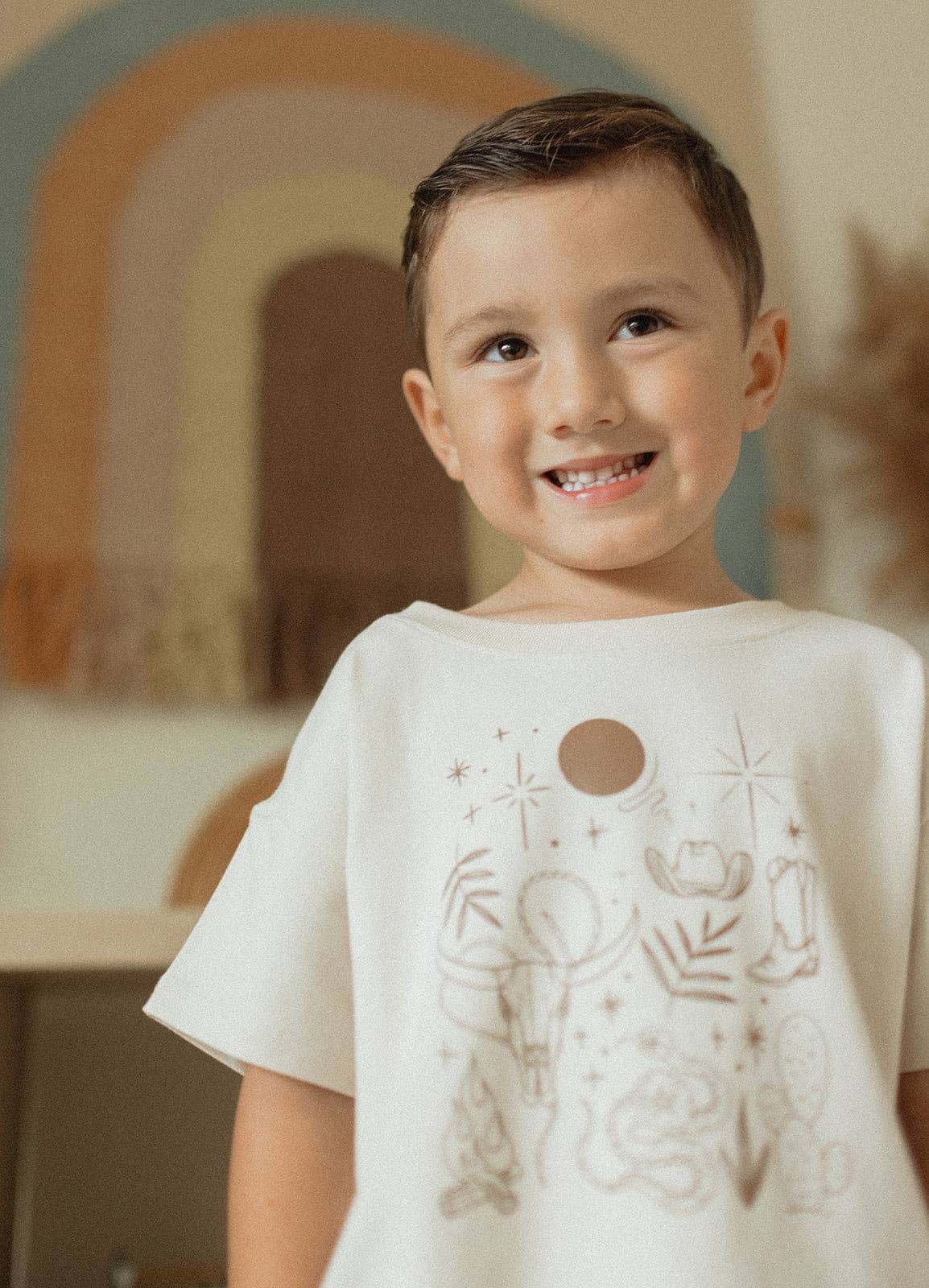 
                  
                    Child wearing a white t-shirt with graphic designs in a softly blurred indoor setting.
                  
                