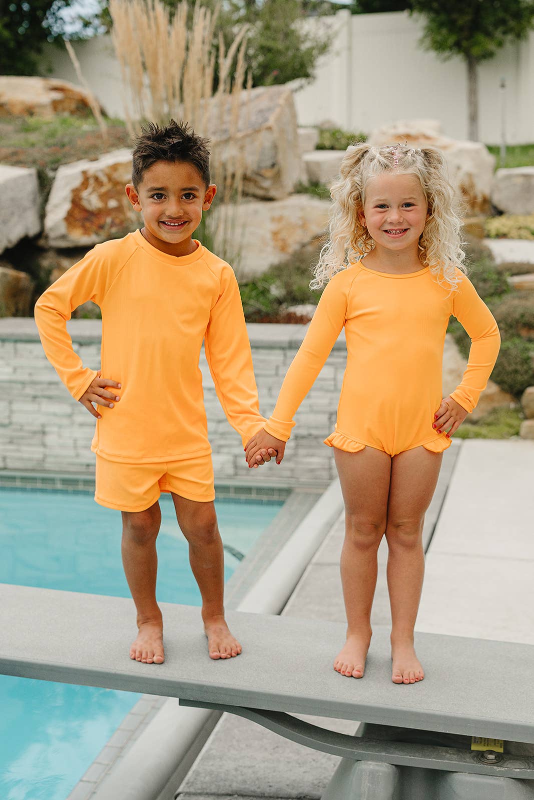 
                  
                    Two children in matching orange swimsuits standing on a pool deck.
                  
                